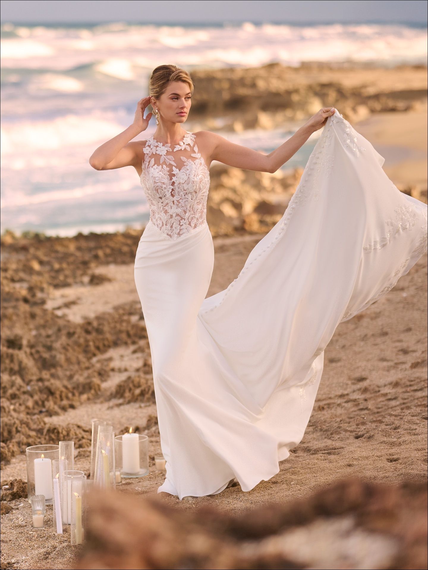 Bride in white wedding dress on beach, holding train, waves in background.