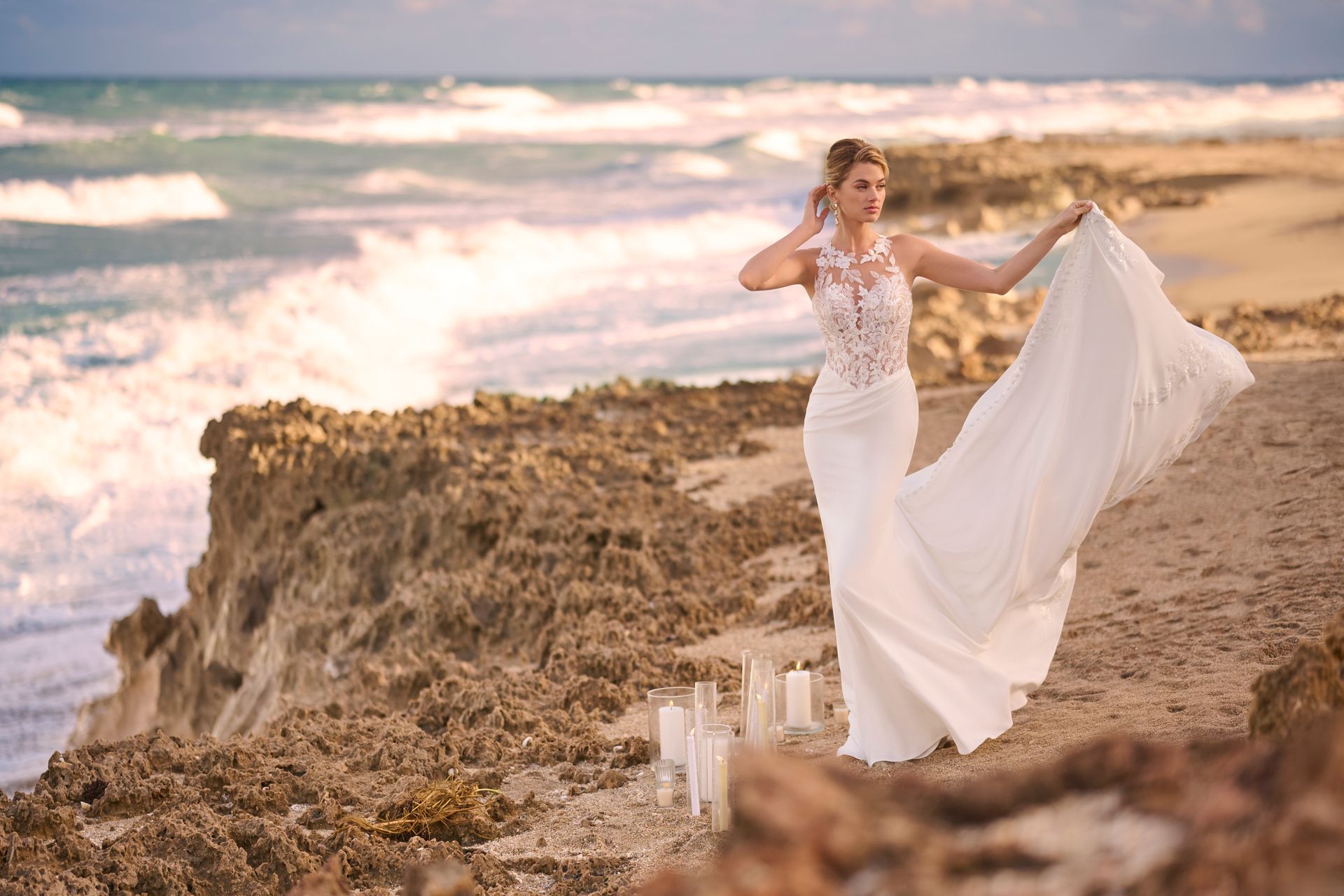Bride in white dress poses on rocky coast, waves in background.