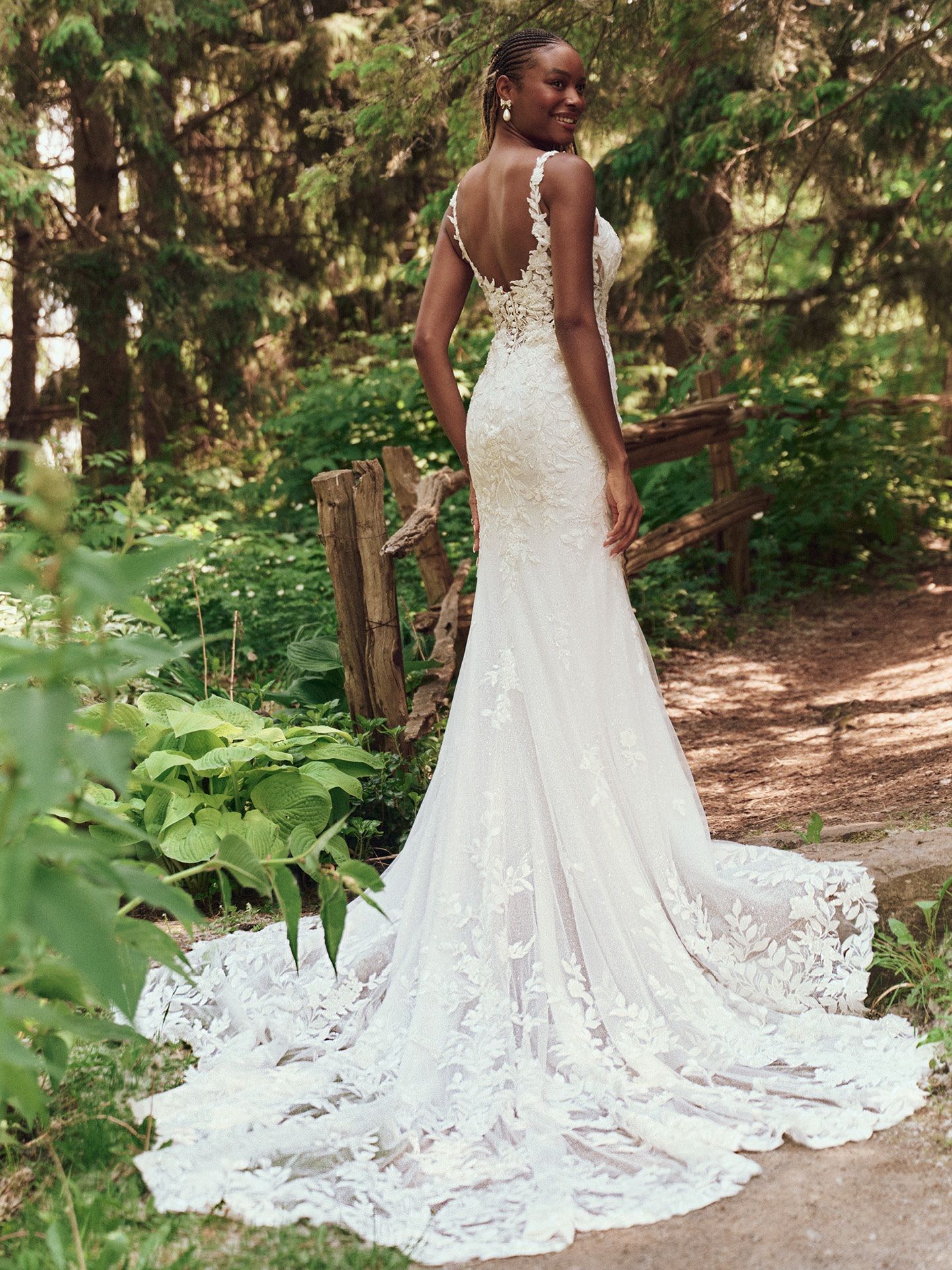 Woman in a lace wedding dress with a long train, standing outside near foliage and a wooden fence.