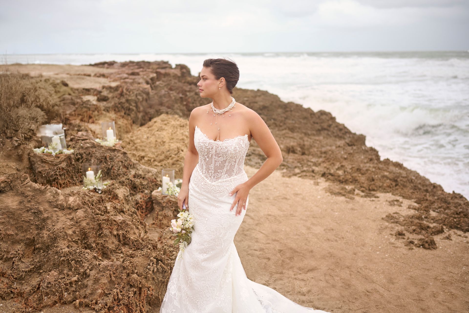 Bride in white lace gown on rocky beach, holding bouquet, looking away from the camera.
