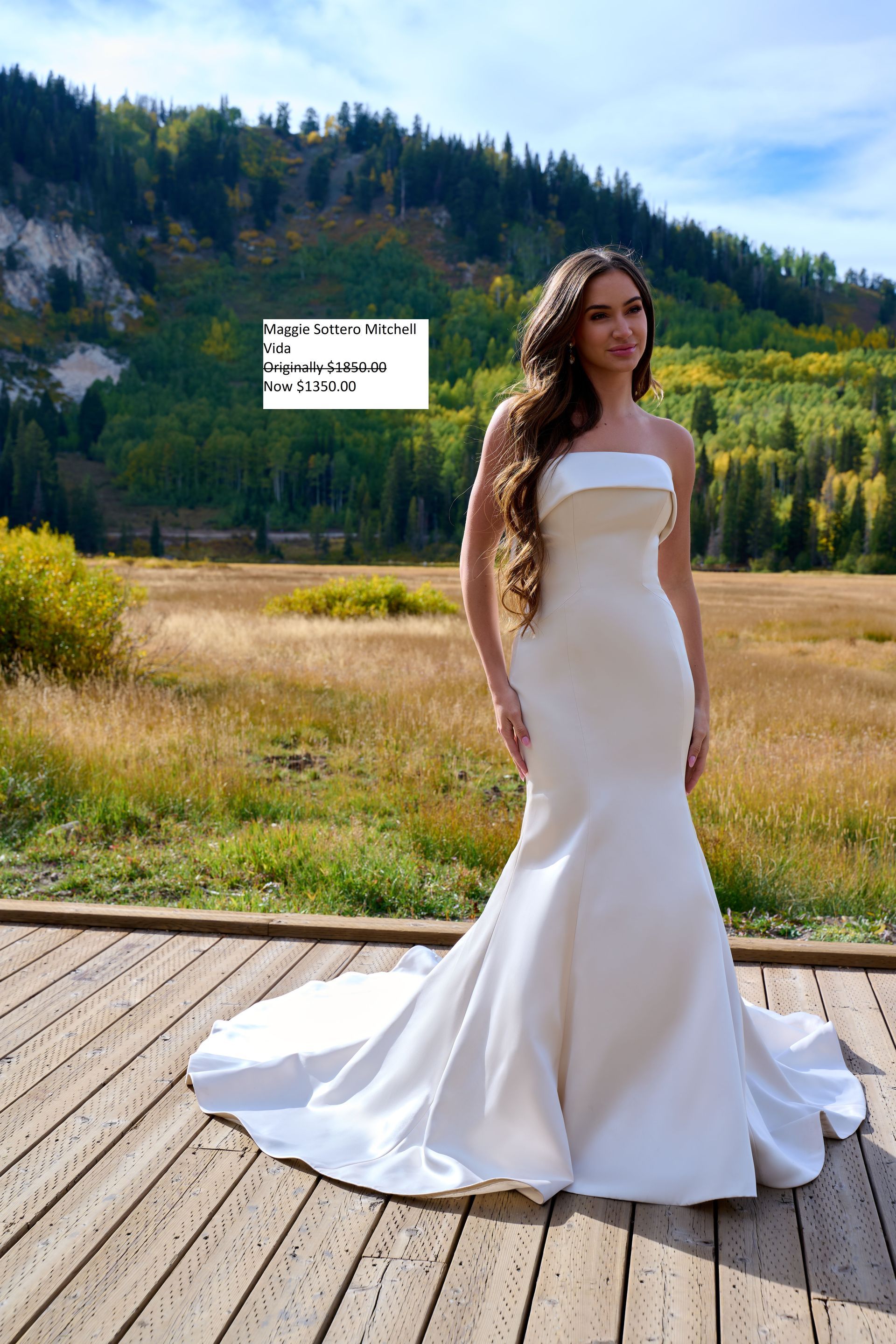 Woman in white strapless wedding dress stands on wooden deck, outdoors with trees and field. Maggie Sottero Mitchell Vida