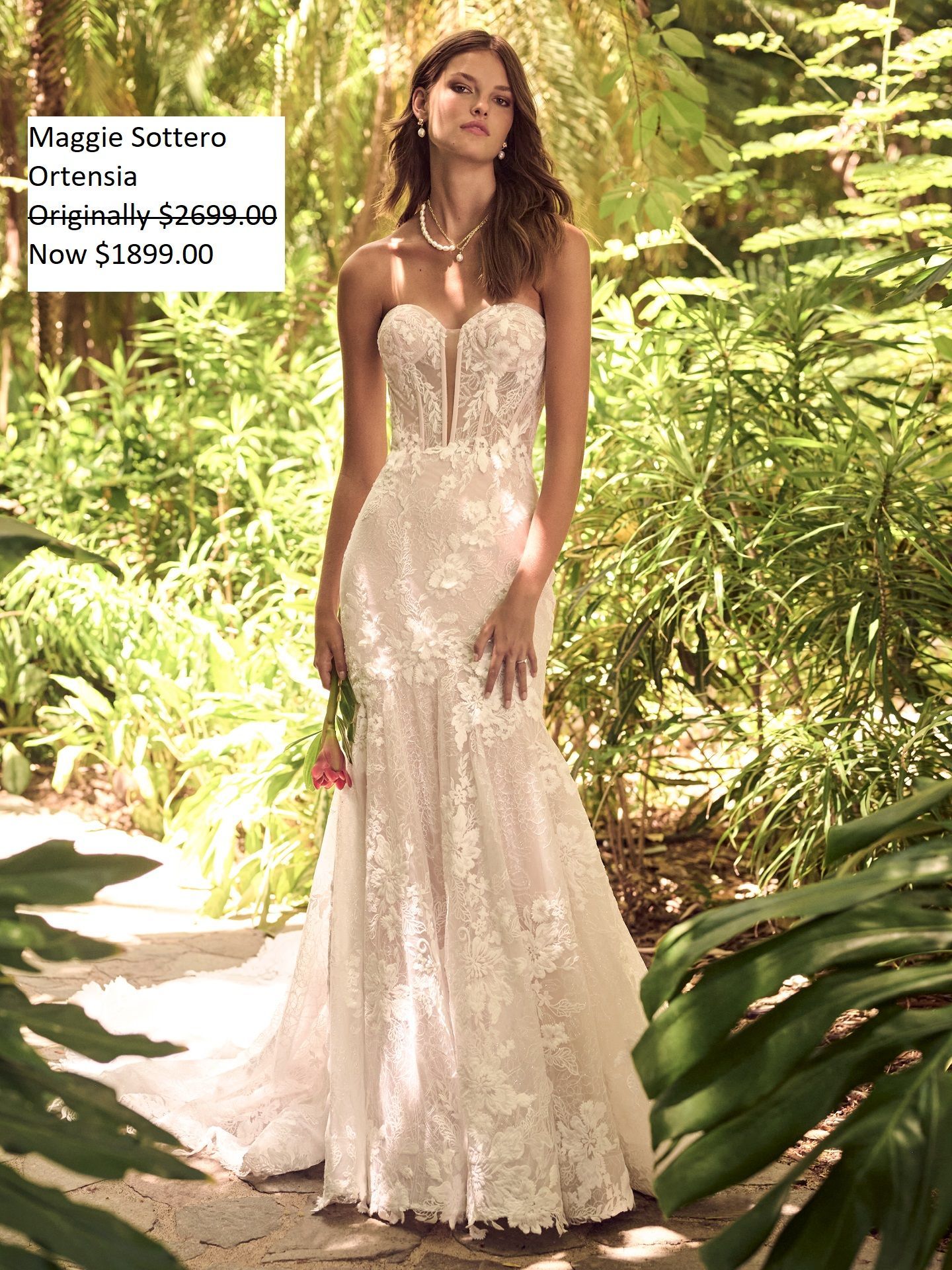 Woman wearing a white lace strapless wedding gown, holding a flower, standing outside. Maggie Sottero Ortensia