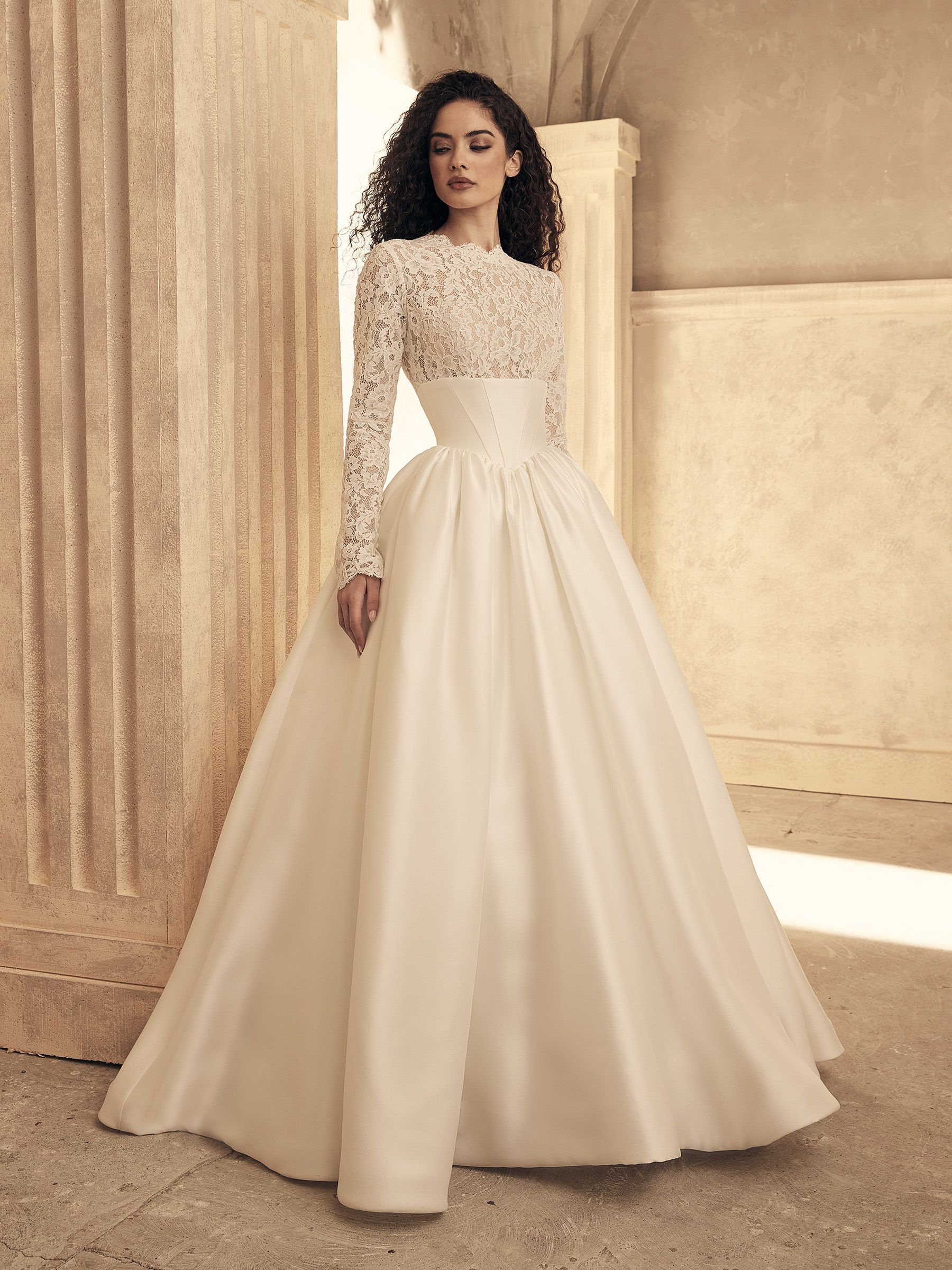 Woman in white wedding dress with lace bodice, long sleeves, and full skirt, standing by a pillar.
