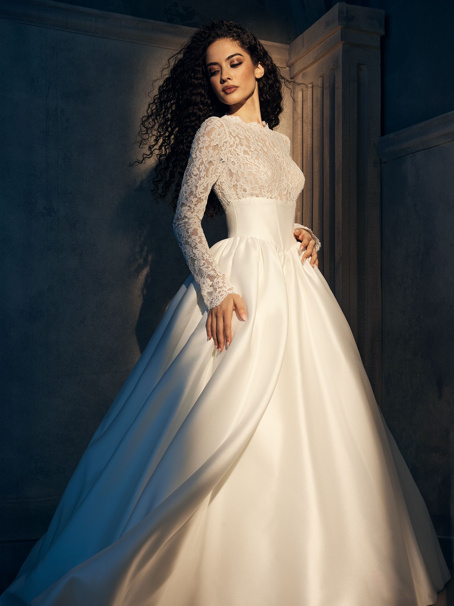 Woman in a white satin wedding gown, lace bodice, curly hair, posing against a wall.