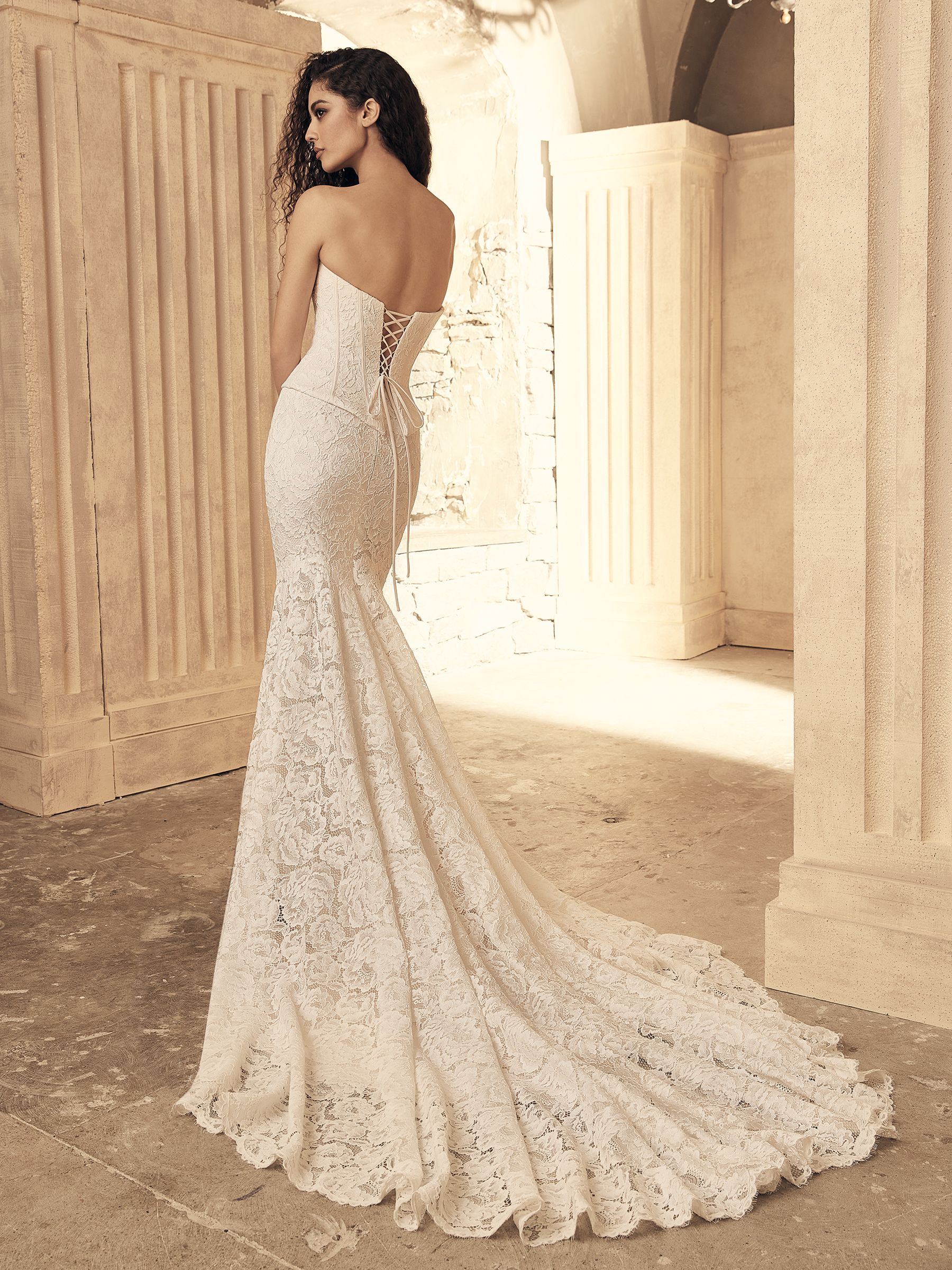 Woman in strapless lace wedding dress with a long train, standing indoors.
