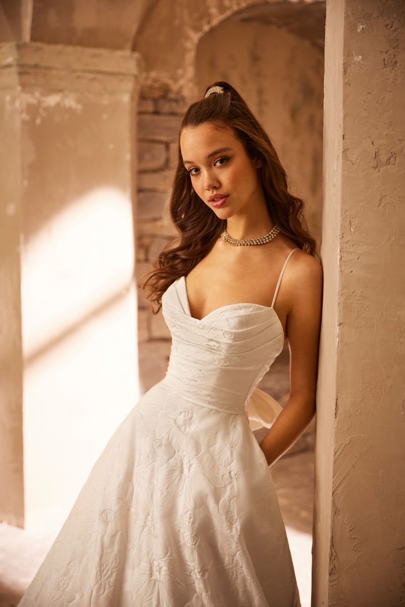 A woman in a white wedding dress is leaning against a wall.