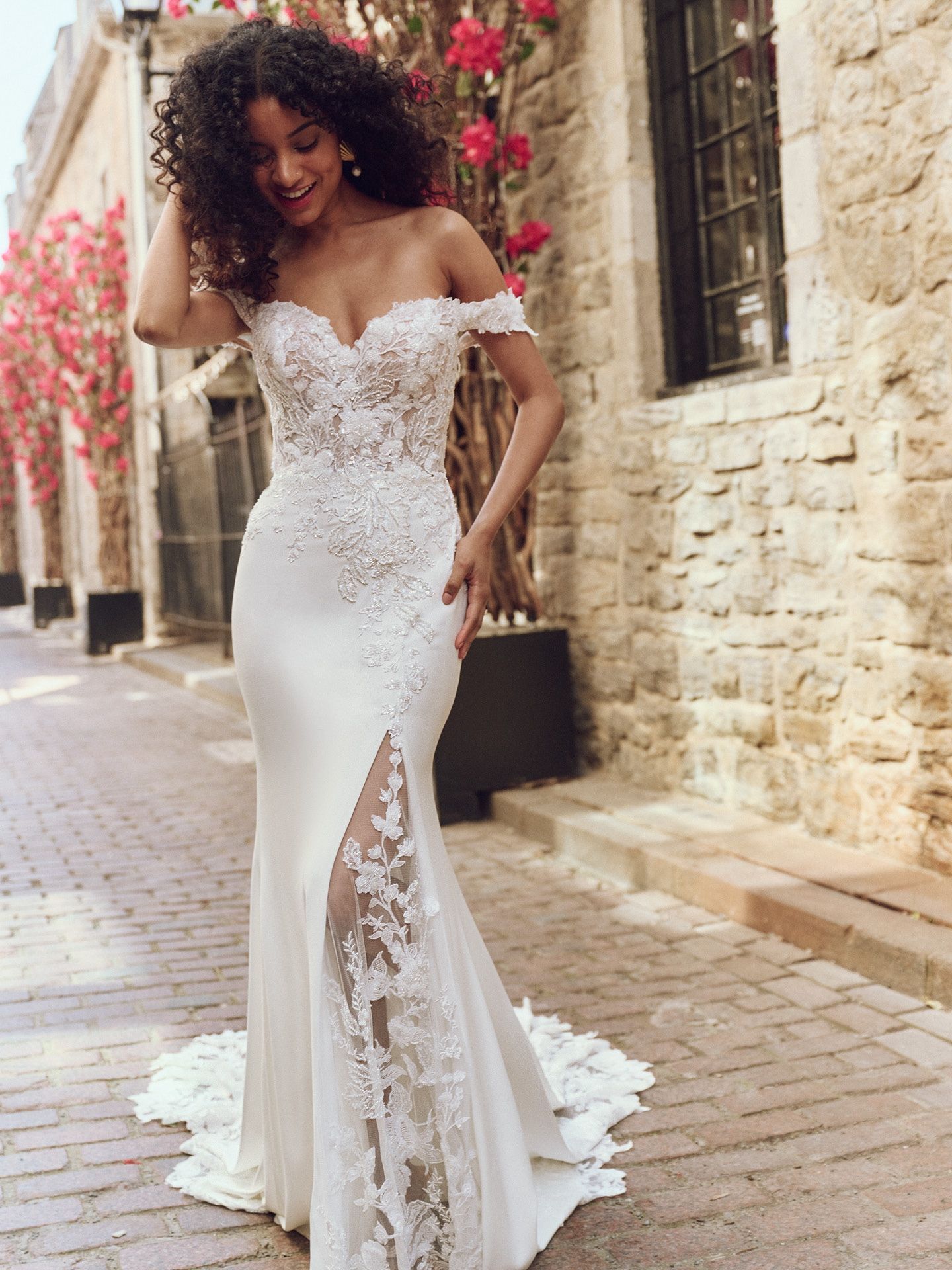 Woman in a white off-the-shoulder wedding dress with a high slit, smiling outside a stone building.