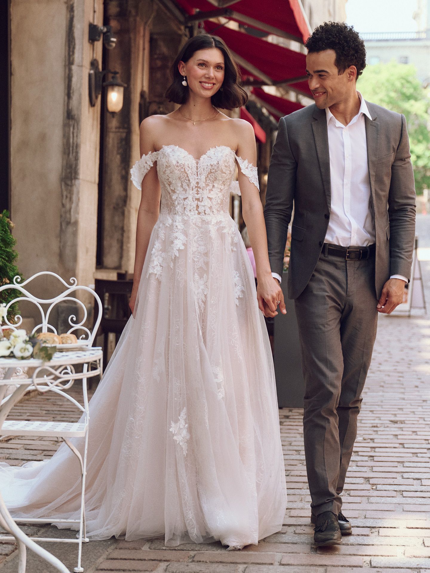 Bride in off-the-shoulder gown and groom in suit walk hand-in-hand down a cobblestone street, smiling.