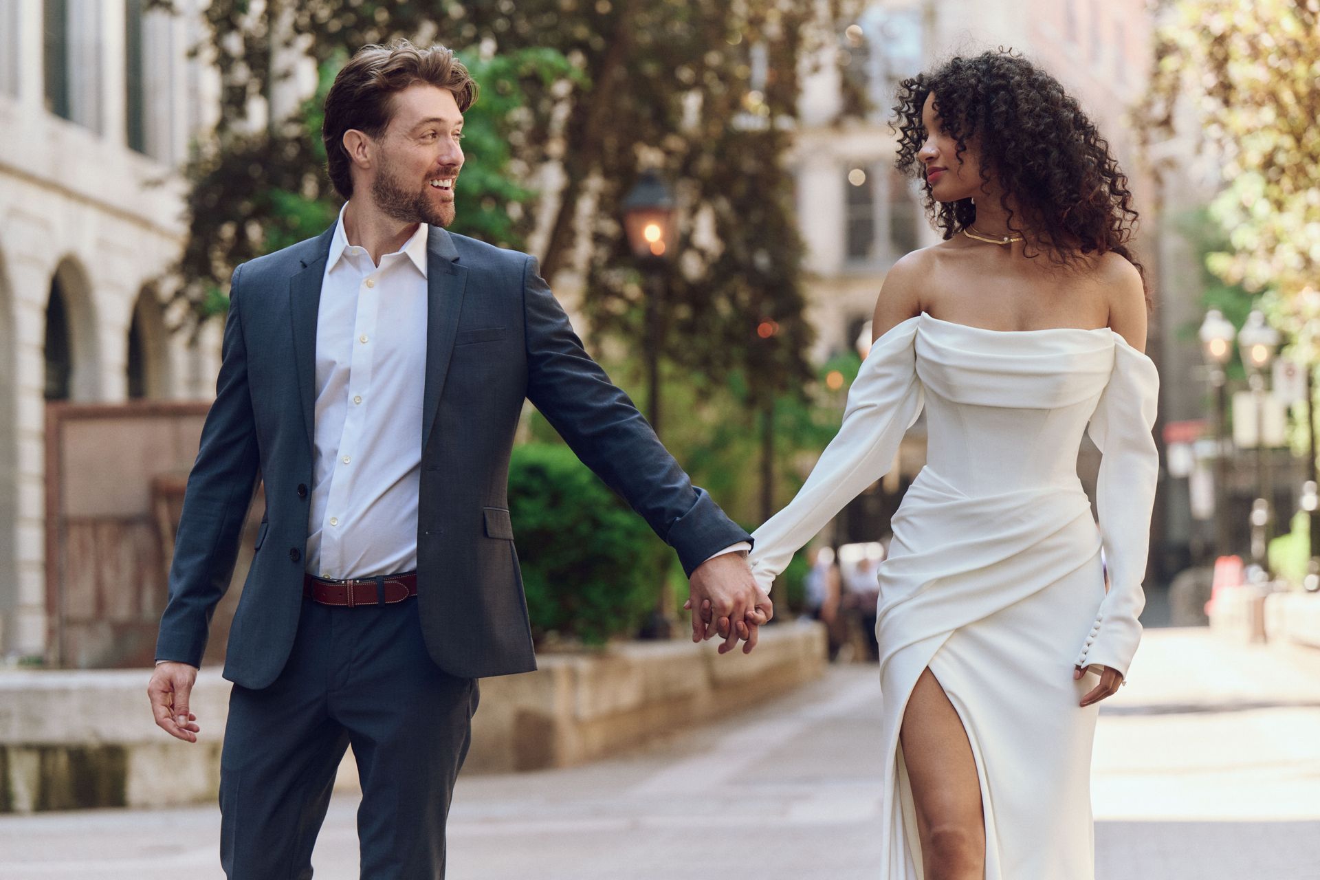 Couple, man in suit and woman in white dress, walking, holding hands, in a city street.