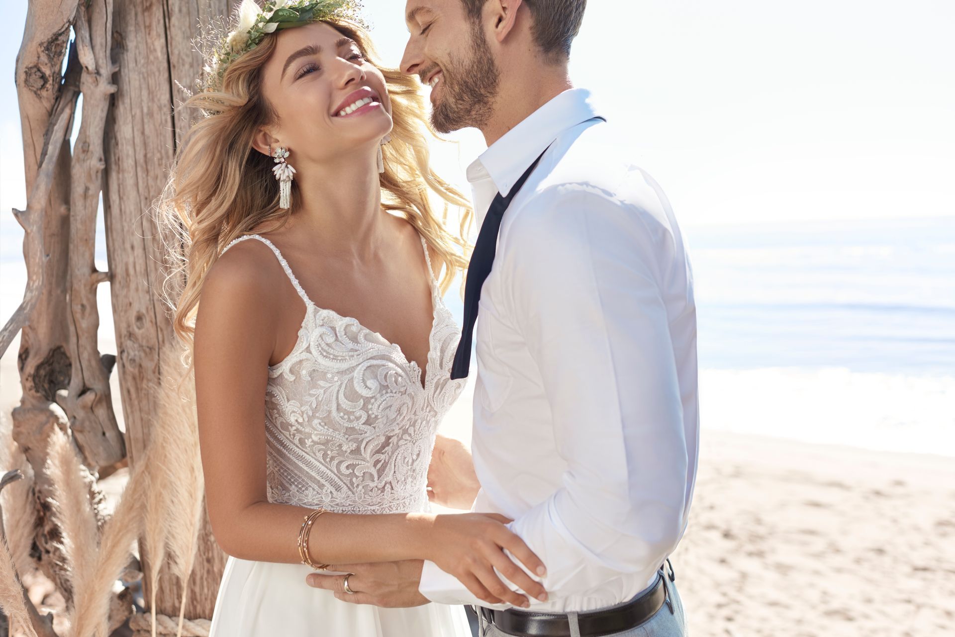 Bride and groom smiling on a beach; she wears a lace wedding dress, he wears a white shirt and tie.