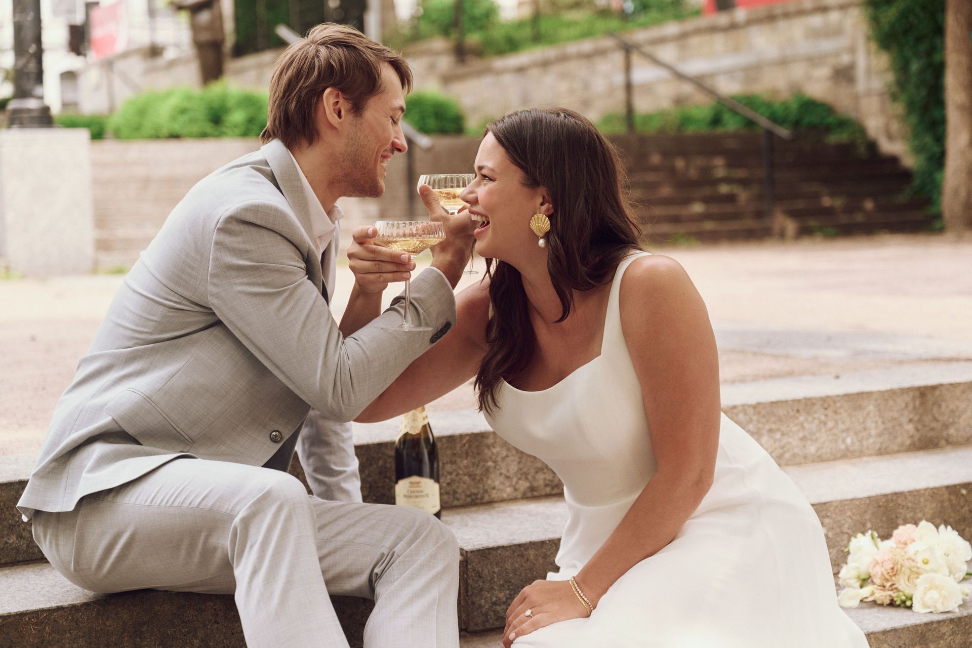 Bride and groom on stone steps share champagne, laughing. The groom wears gray, the bride white; setting appears urban.