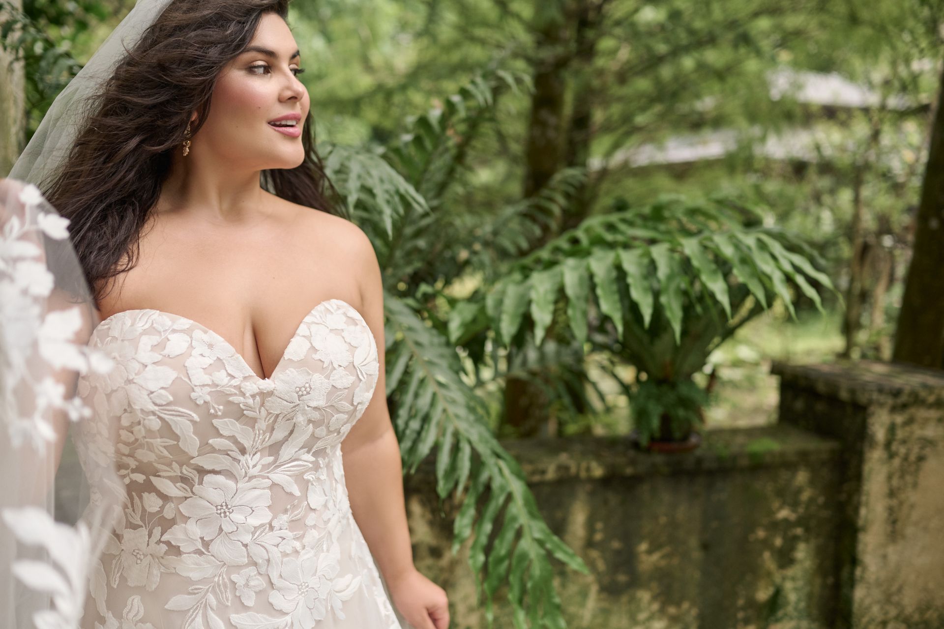 Bride in a strapless lace wedding dress, smiling outdoors with lush greenery.
