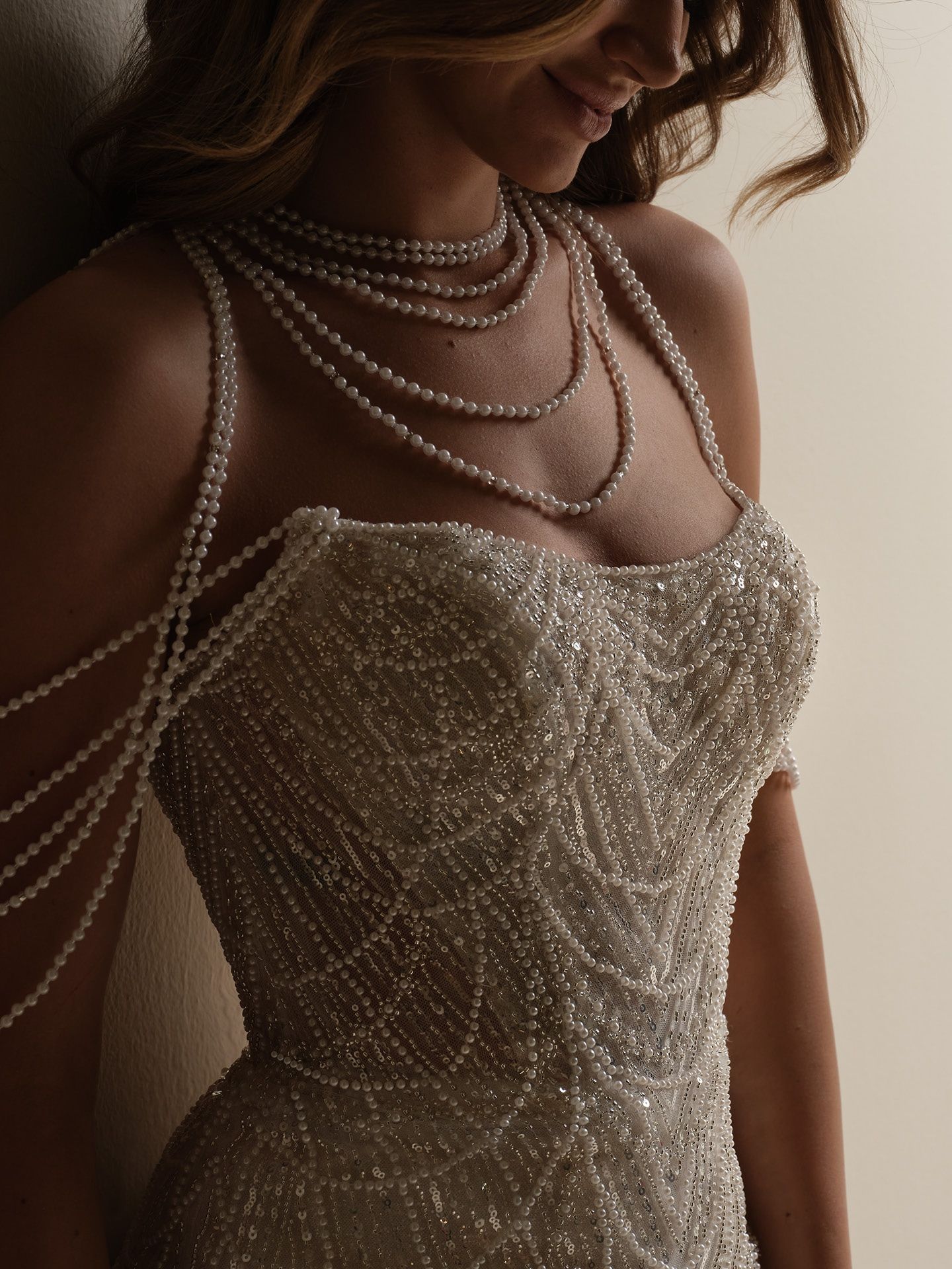 Woman in a shimmering beaded gown, wearing multiple pearl necklaces, smiles gently.