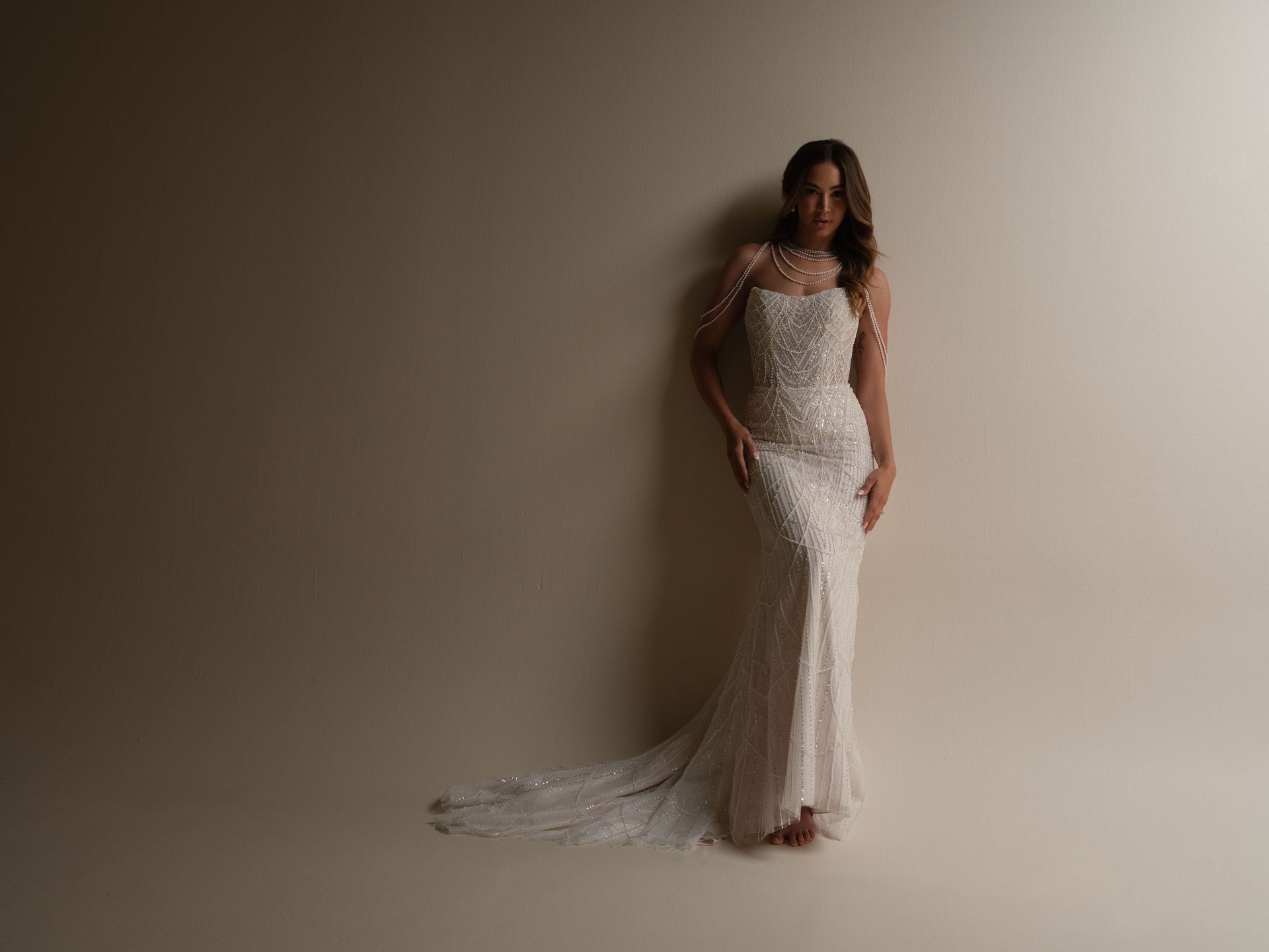 Woman in a white beaded gown, leaning against a neutral wall.