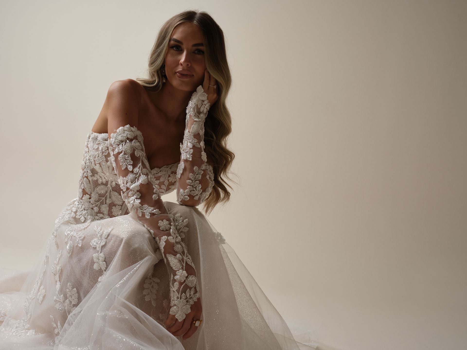 Woman in a white, off-the-shoulder wedding gown, poses indoors.