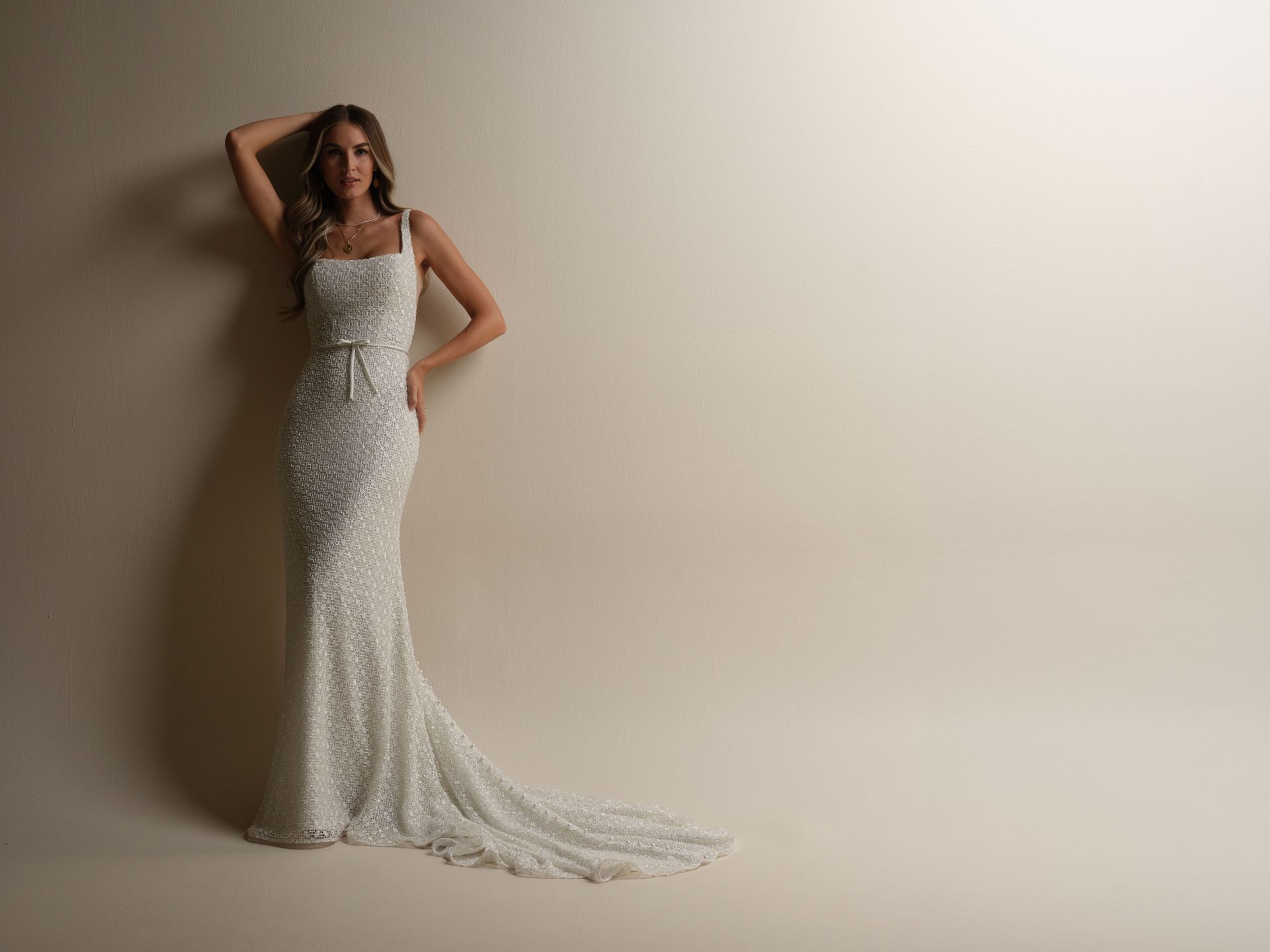 Woman in a beaded white gown poses against a beige wall, hand on head, train flowing.