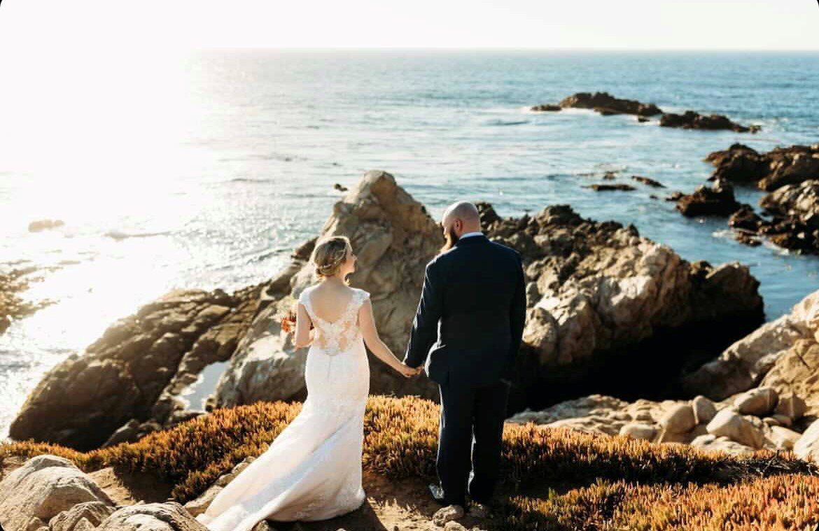 A bride and groom are holding hands while standing on a rocky cliff overlooking the ocean.