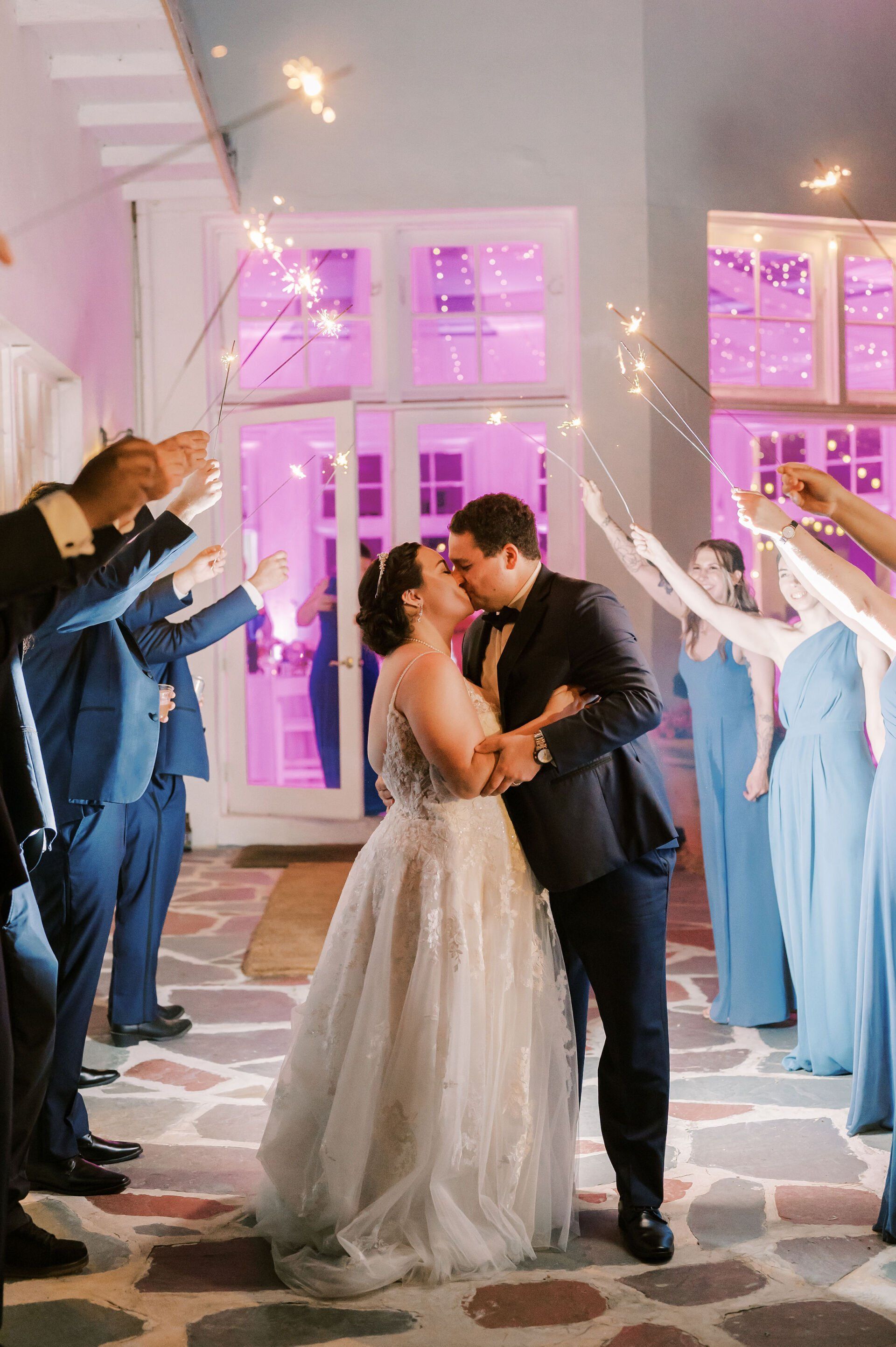 A bride and groom kissing while their wedding party holds sparklers in the air.