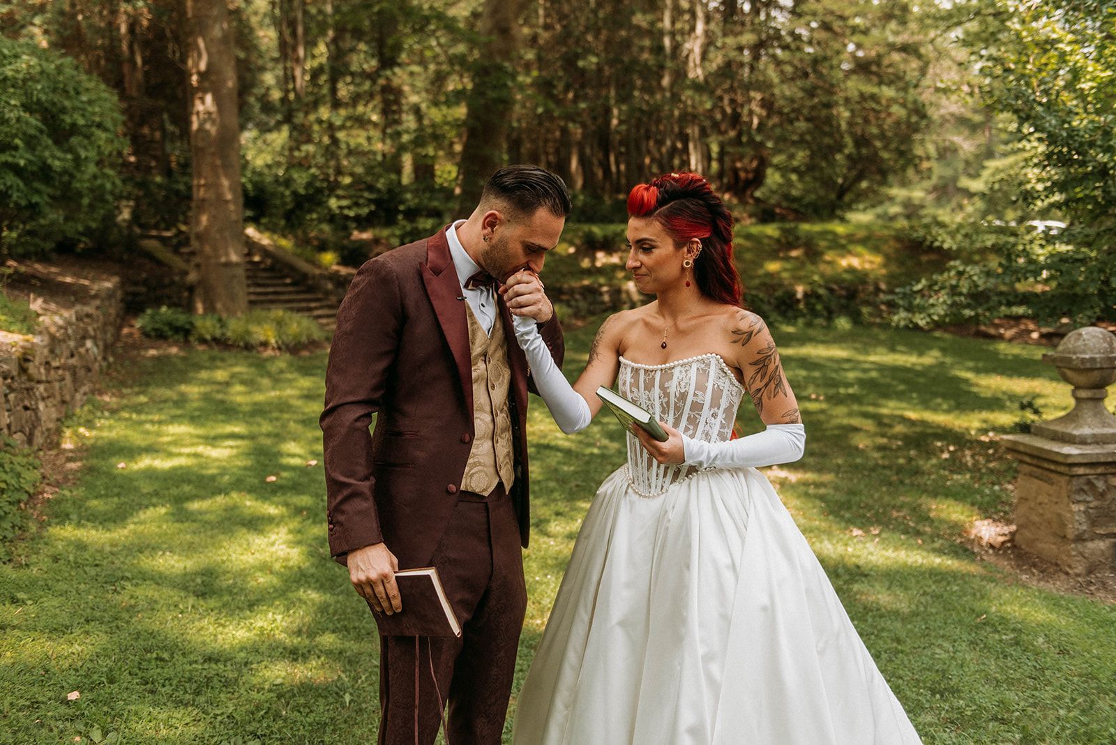 Groom kisses bride's hand in garden. Bride in white dress with arm tattoos. Groom in burgundy suit.
