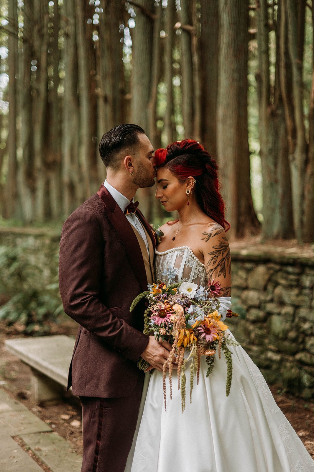 Bride and groom in burgundy suit and white gown, embrace in forest setting. Man kisses woman's forehead.