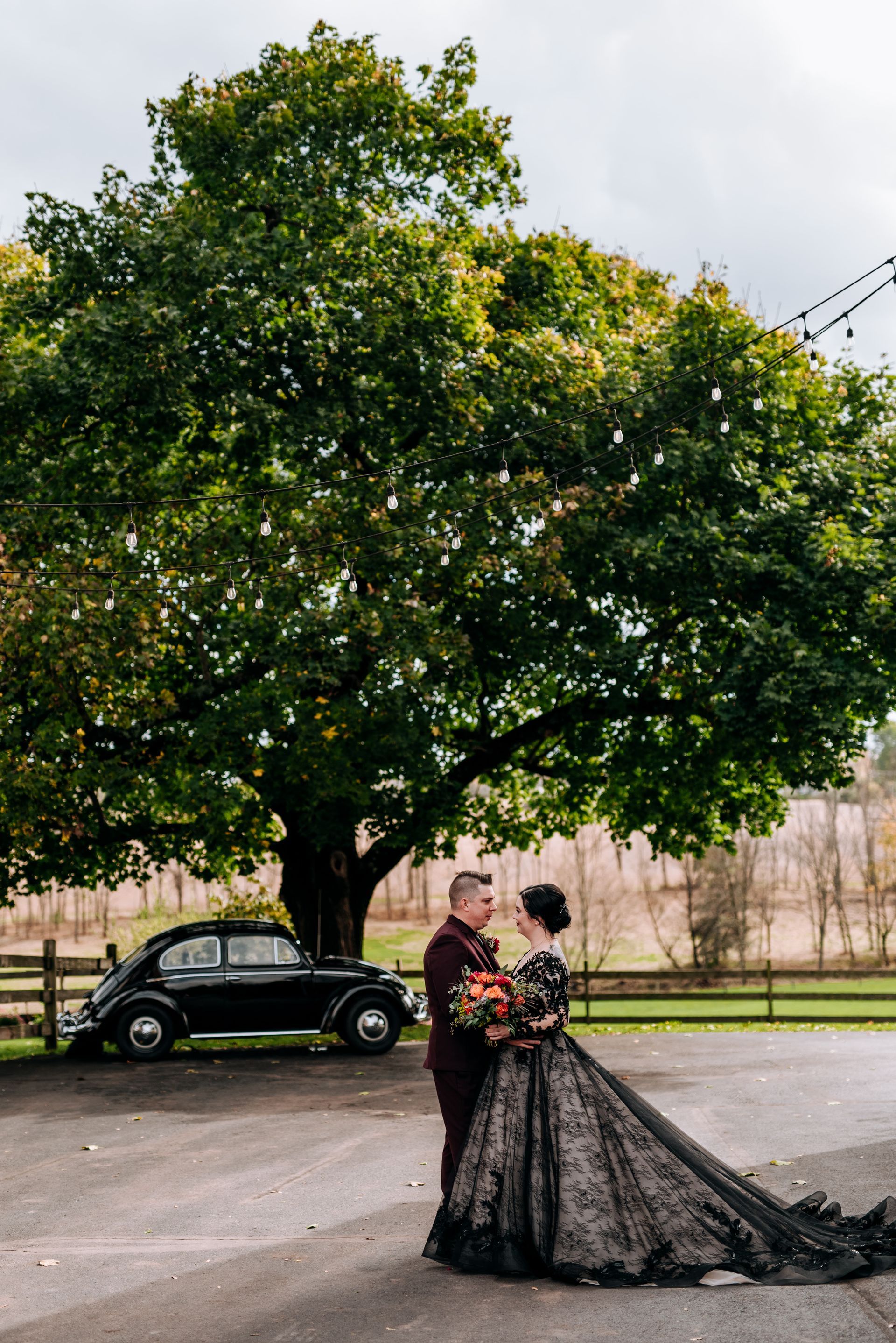 Wedding couple in black attire under a large tree, black car in the background.