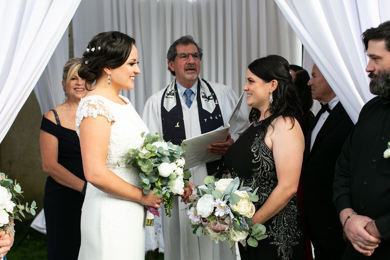 Two women in wedding attire at an outdoor ceremony, facing each other. A clergyman officiates.