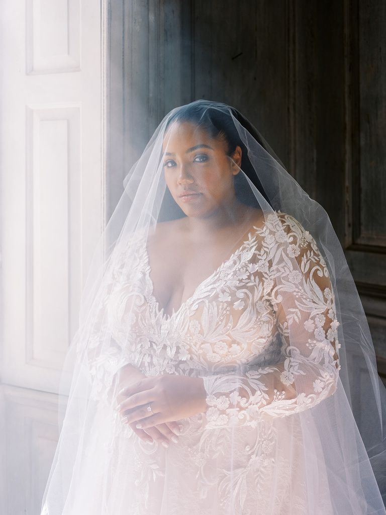 A woman in a wedding dress and veil is standing in front of a window.