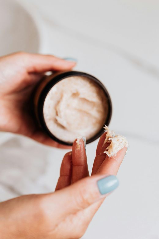 Hands holding jar of skincare product, a finger has a small scoop of cream. Light blue nails.