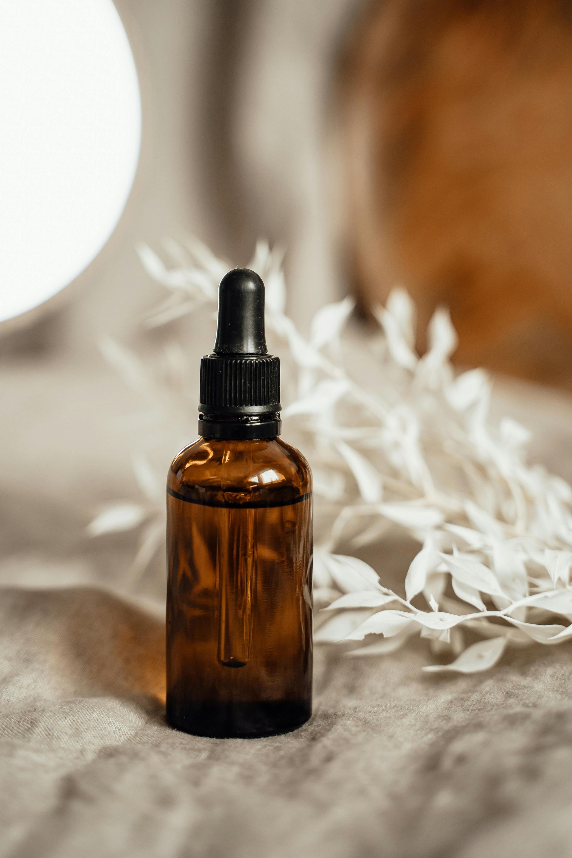 Amber glass bottle with dropper, on a textured surface, with white foliage, soft neutral tones.