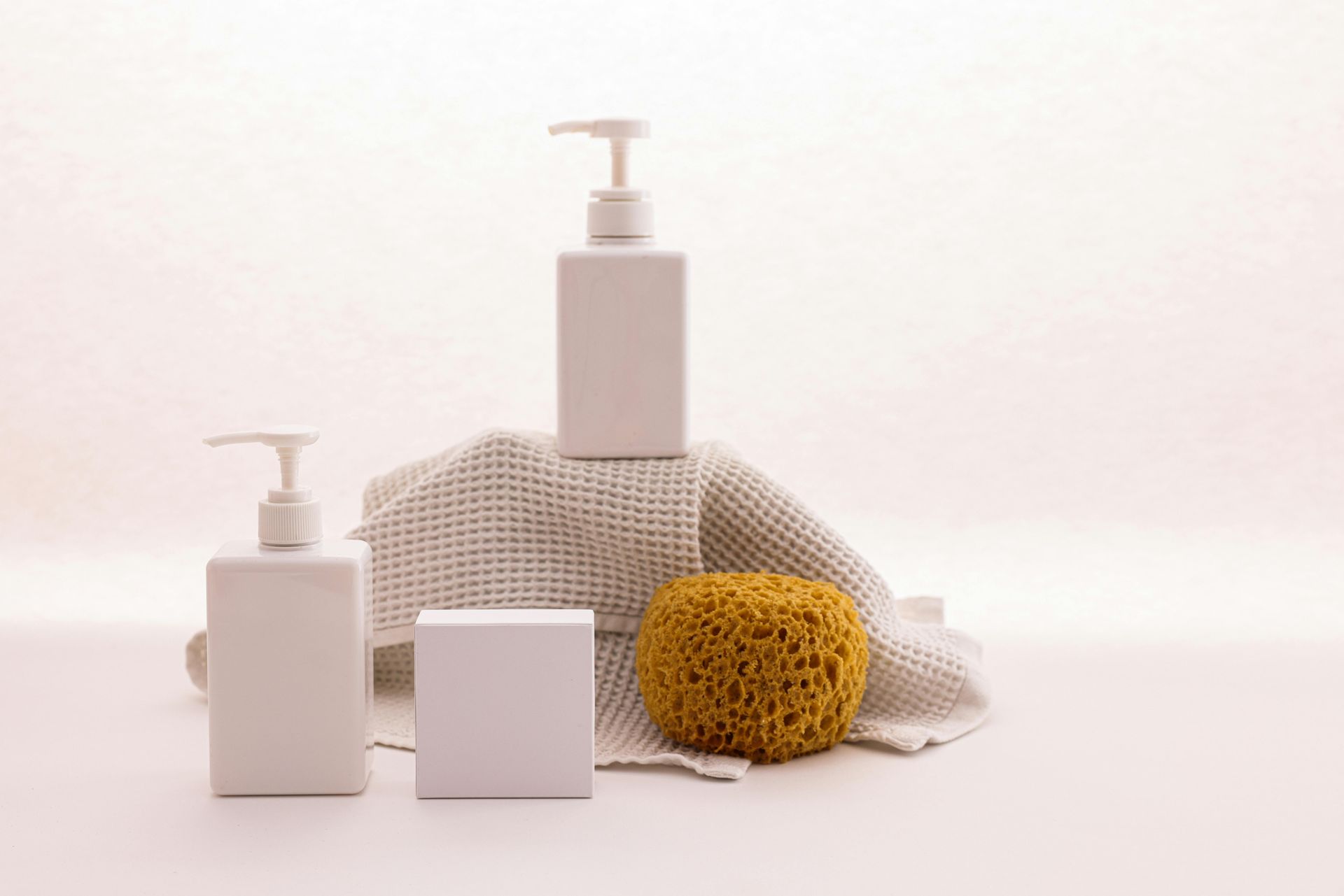 White soap bottles, block, and sponge on a textured cloth against a soft background.