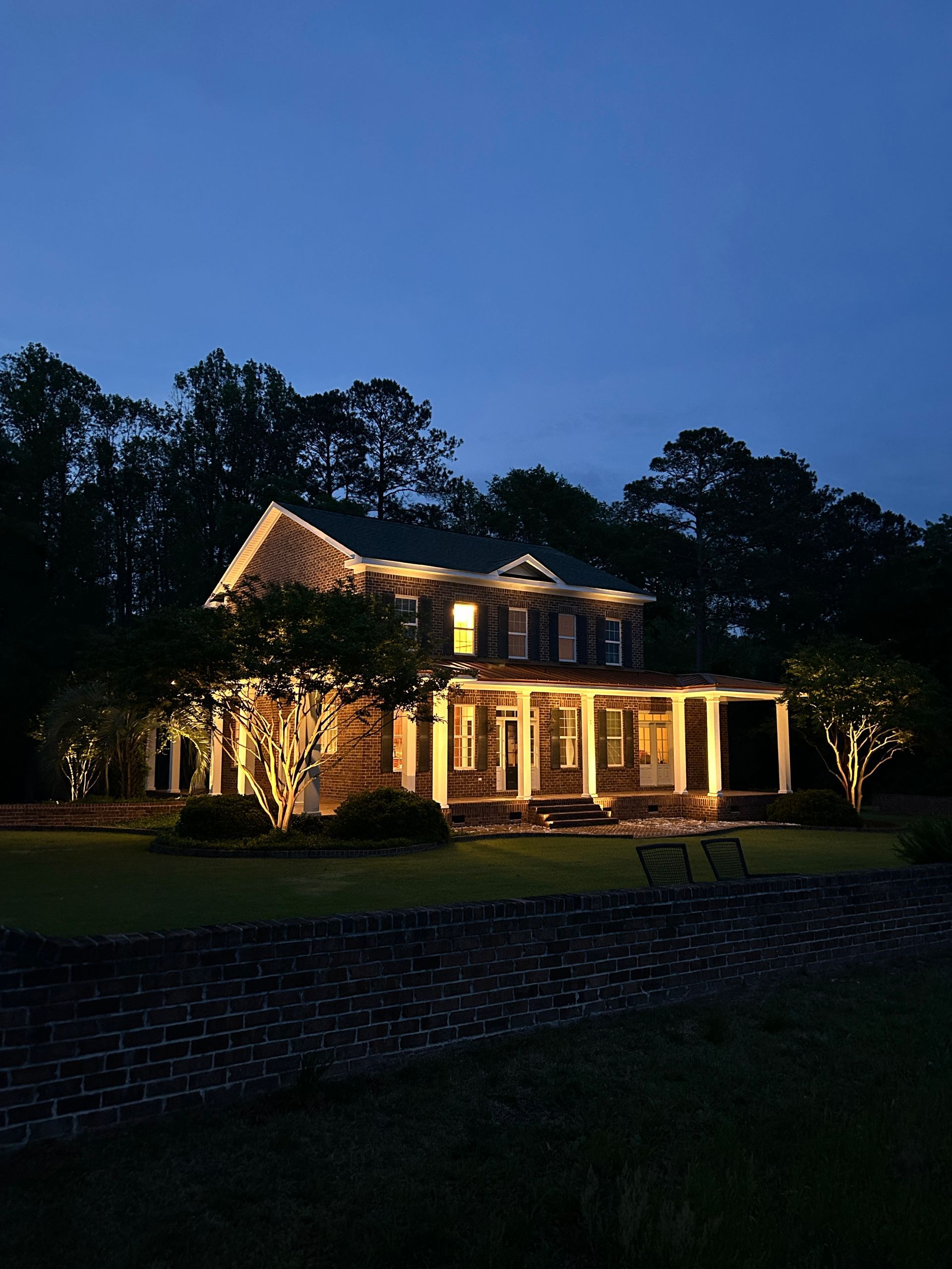 Two-story house illuminated at night with a porch, trees, and a brick wall.