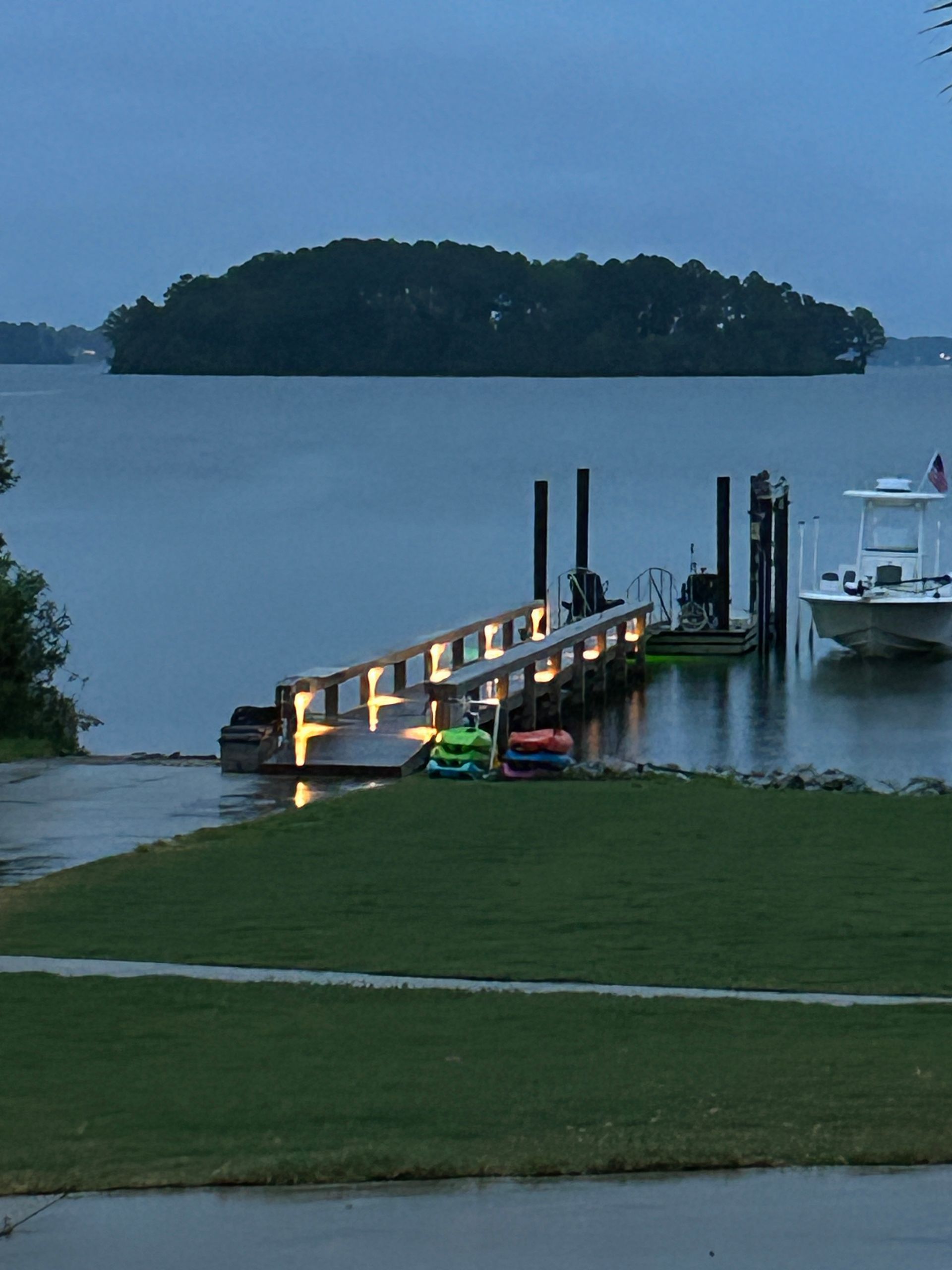 Dock lit up at dusk, overlooking a lake with an island in the distance. Boats are docked nearby.