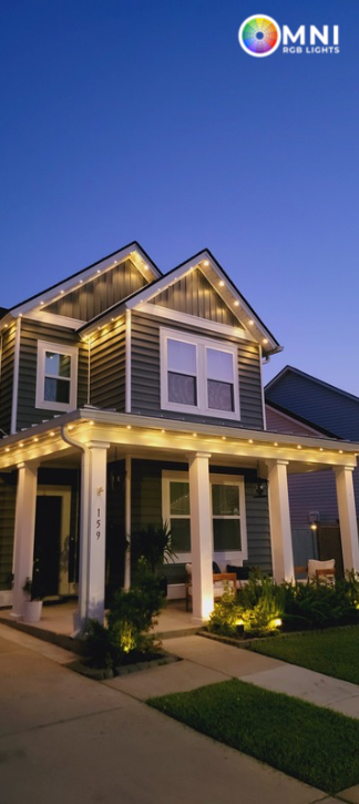 Two-story house with outdoor lighting at dusk. Porch lit, warm lights along roof line. Dark blue sky.