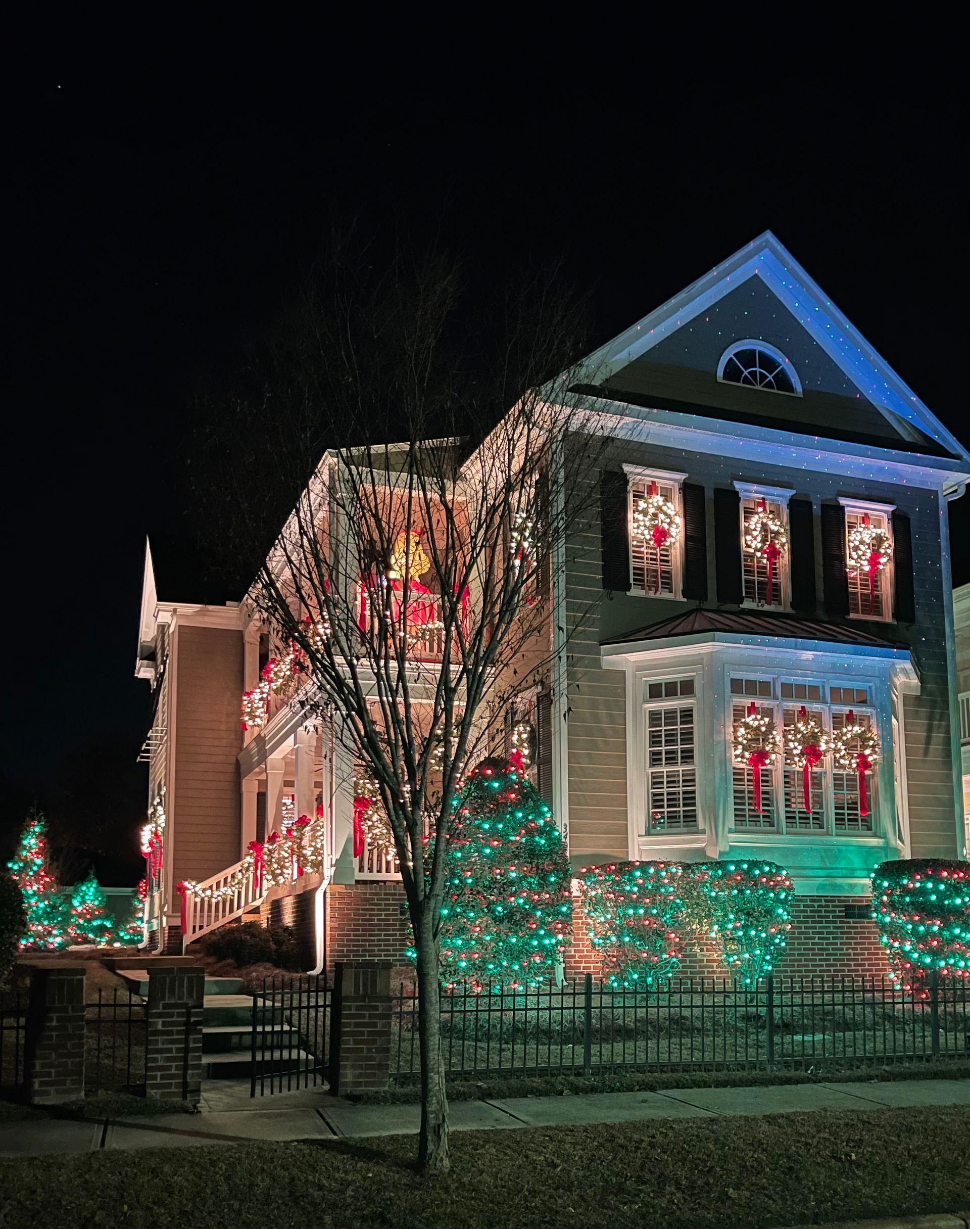 House lit with Christmas lights; green and white lights on bushes, red bows in windows.