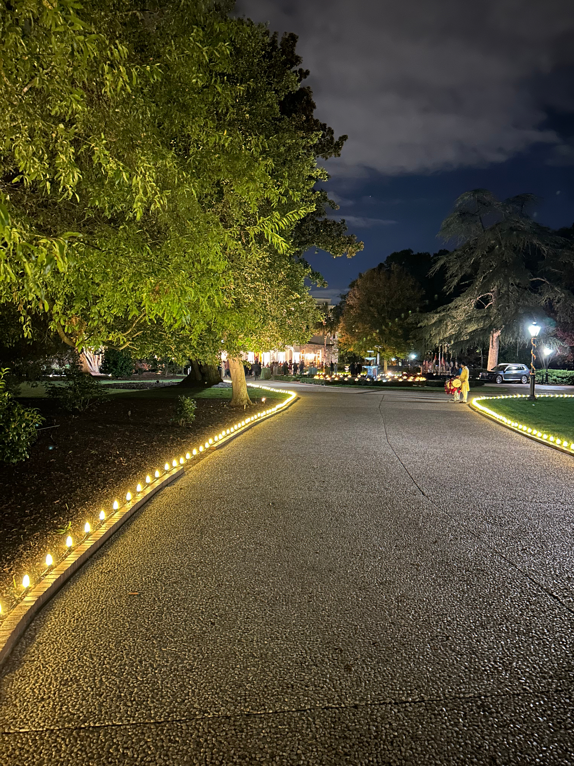 Gravel path lined with lights leads to a building at night. Large trees and a cloudy sky frame the scene.