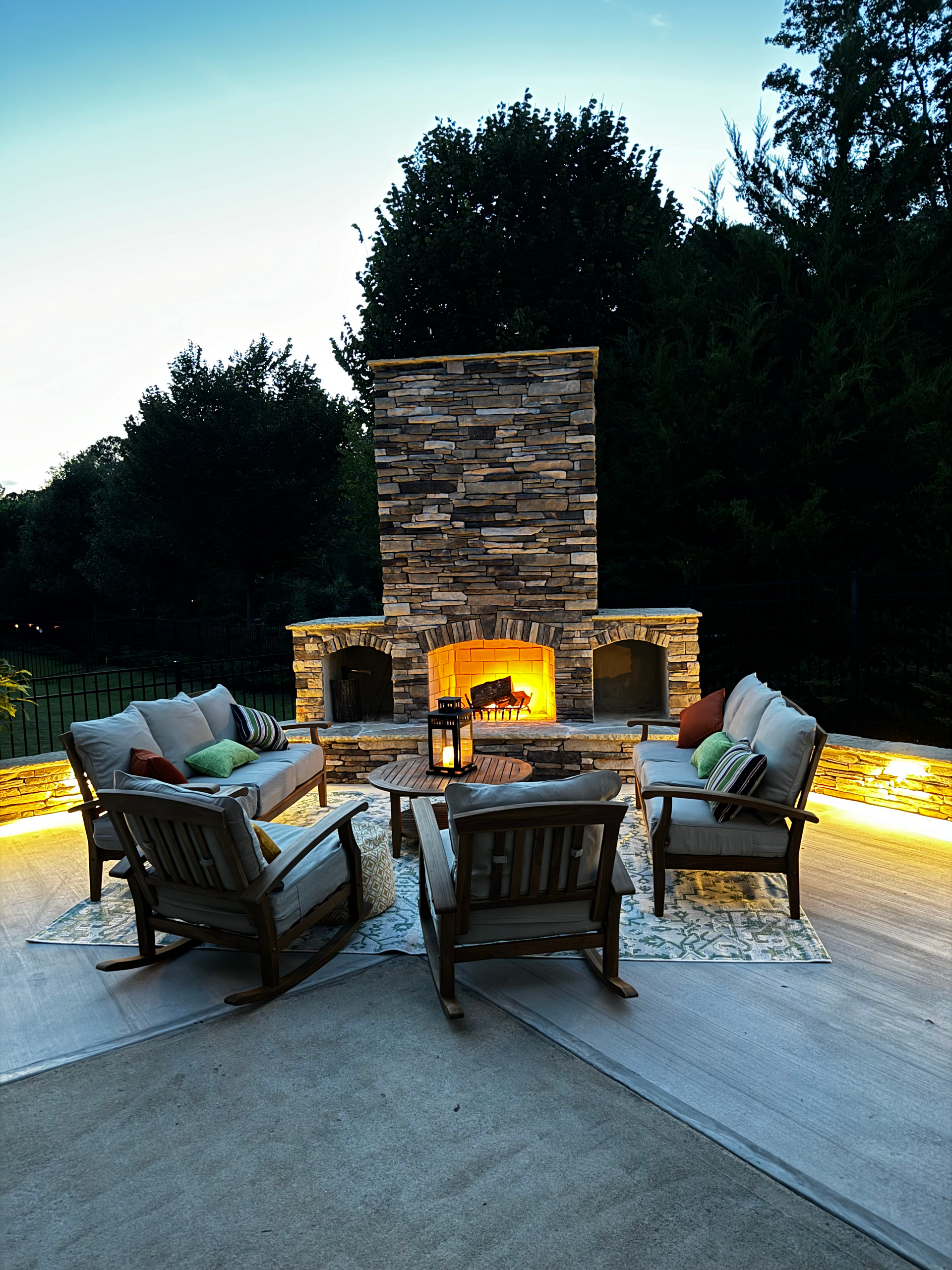 Outdoor stone fireplace with seating, glowing fire, and patio lighting.