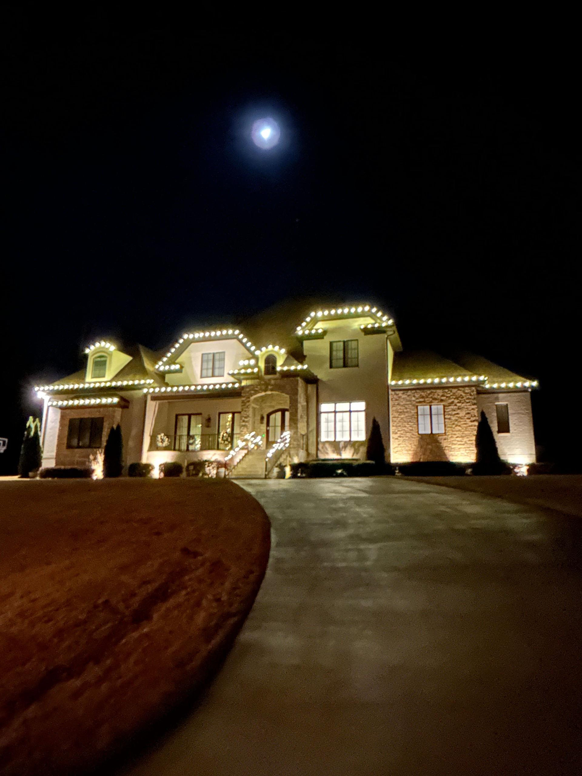 Large house at night, lit with white lights. Dark sky, full moon, long driveway.