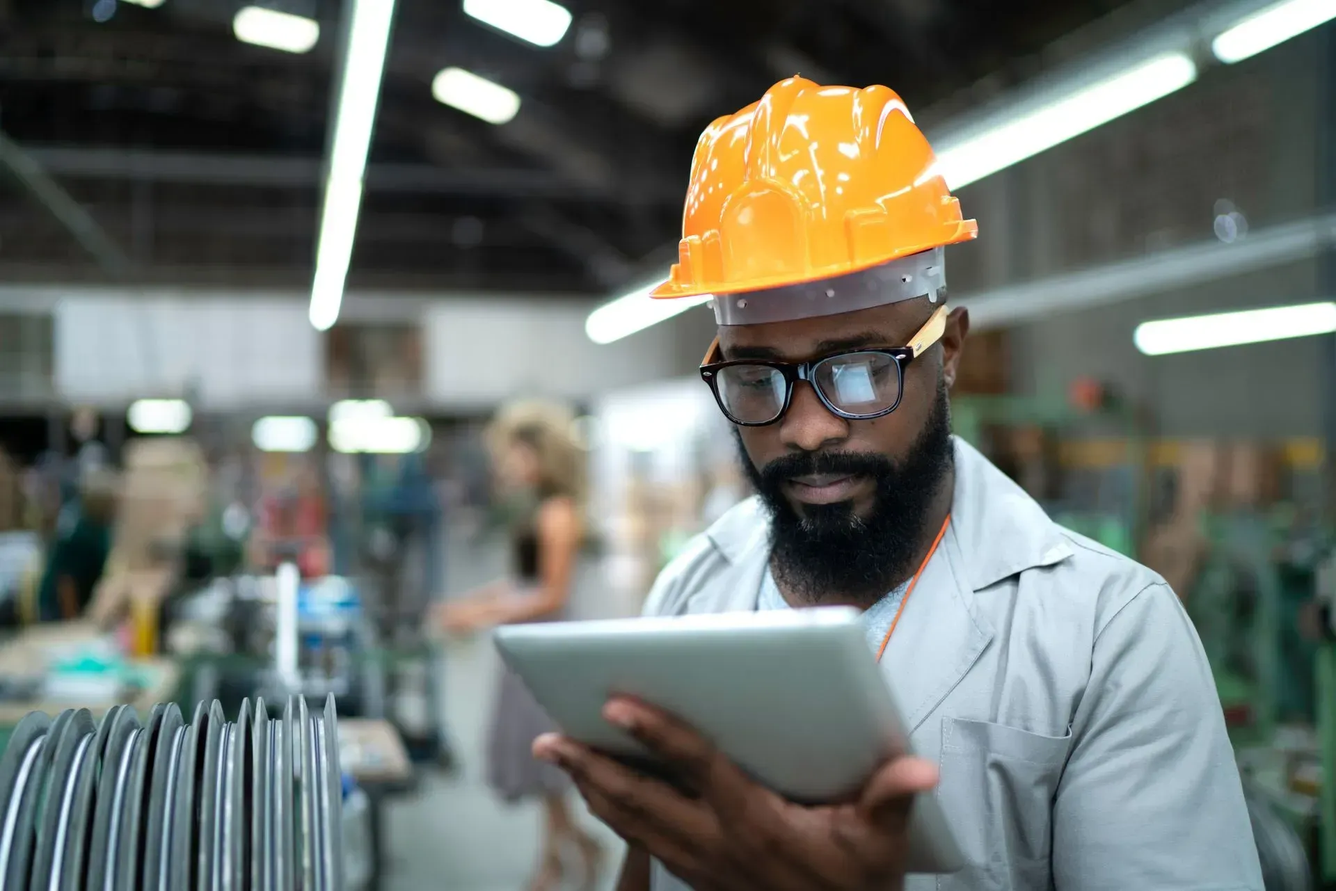 A man wearing a hard hat and glasses is using a tablet in a factory.
