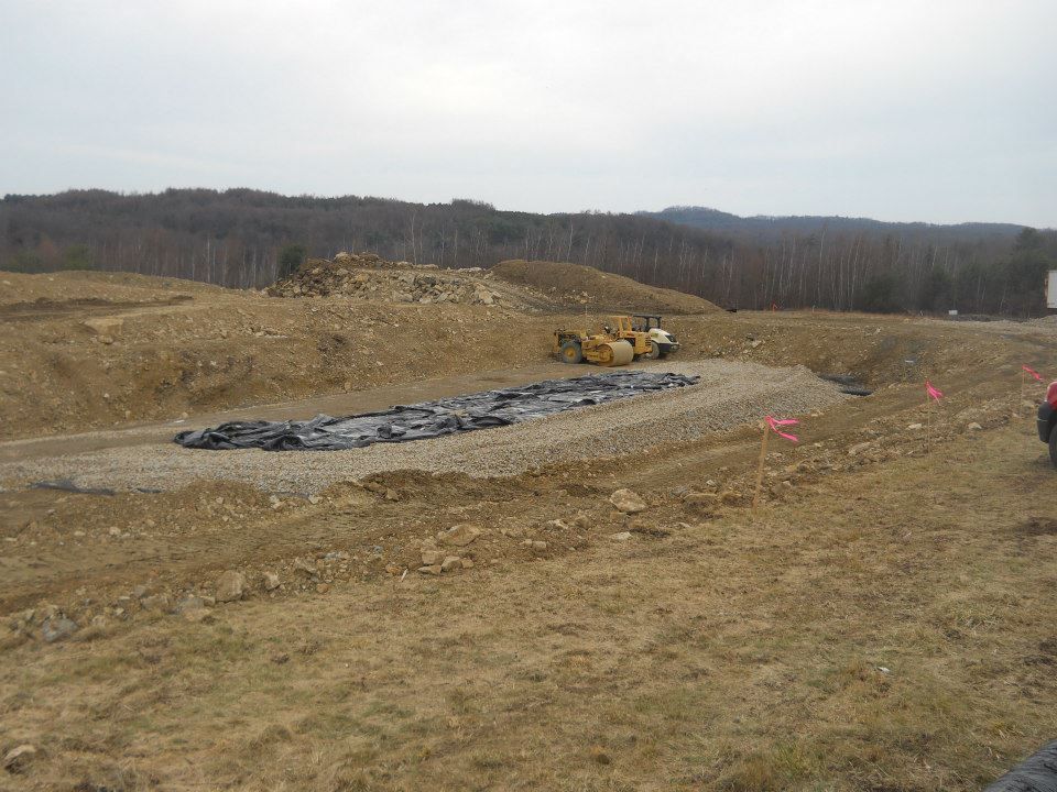 Construction site with a bulldozer, gravel, and a dark layer of material, with a wooded hill in the background.