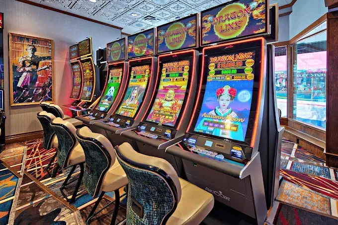Row of slot machines with lit screens in a casino, with chairs in front.