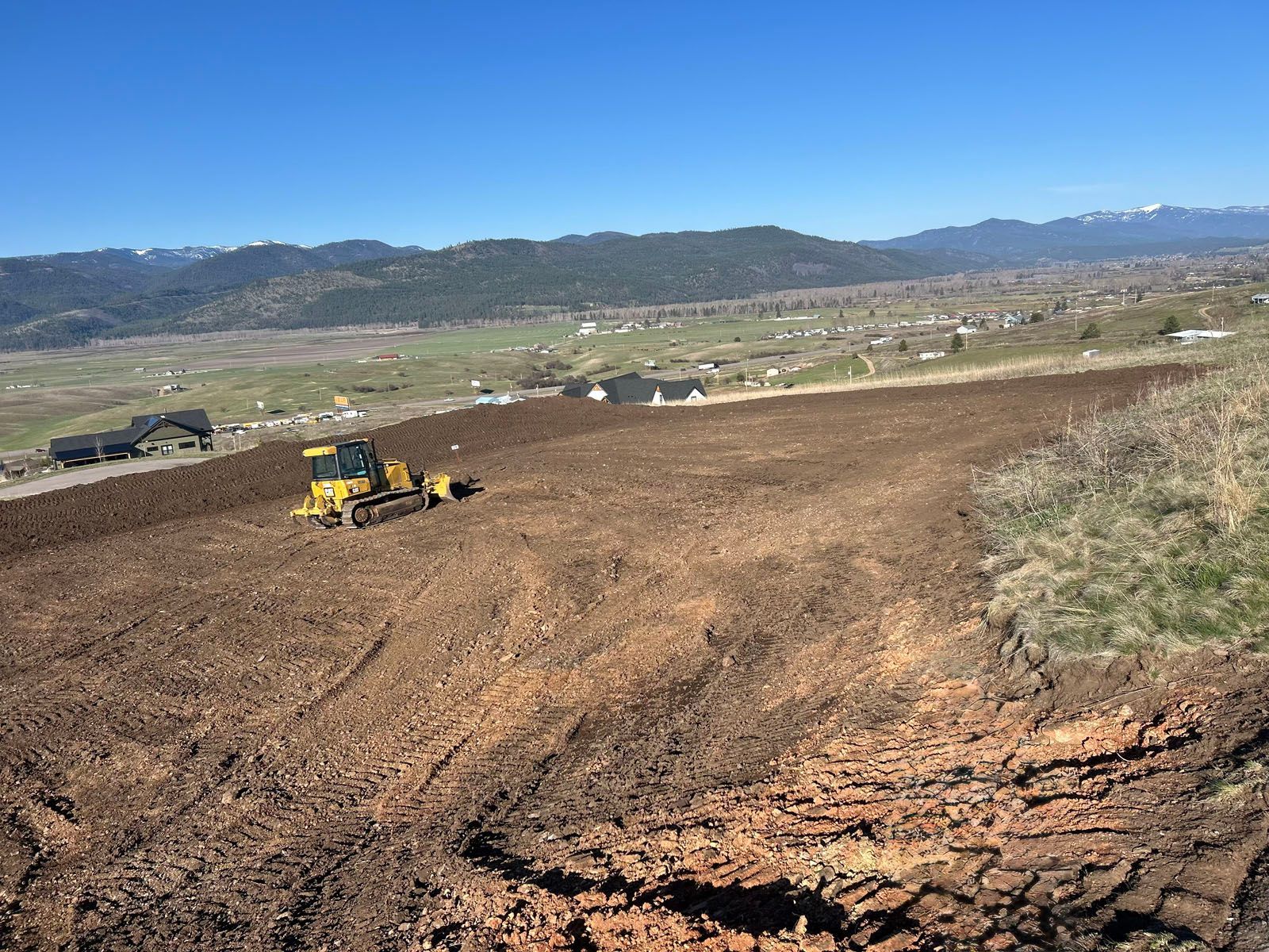 Bulldozer leveling a dirt field with houses and mountains in the background under a blue sky.