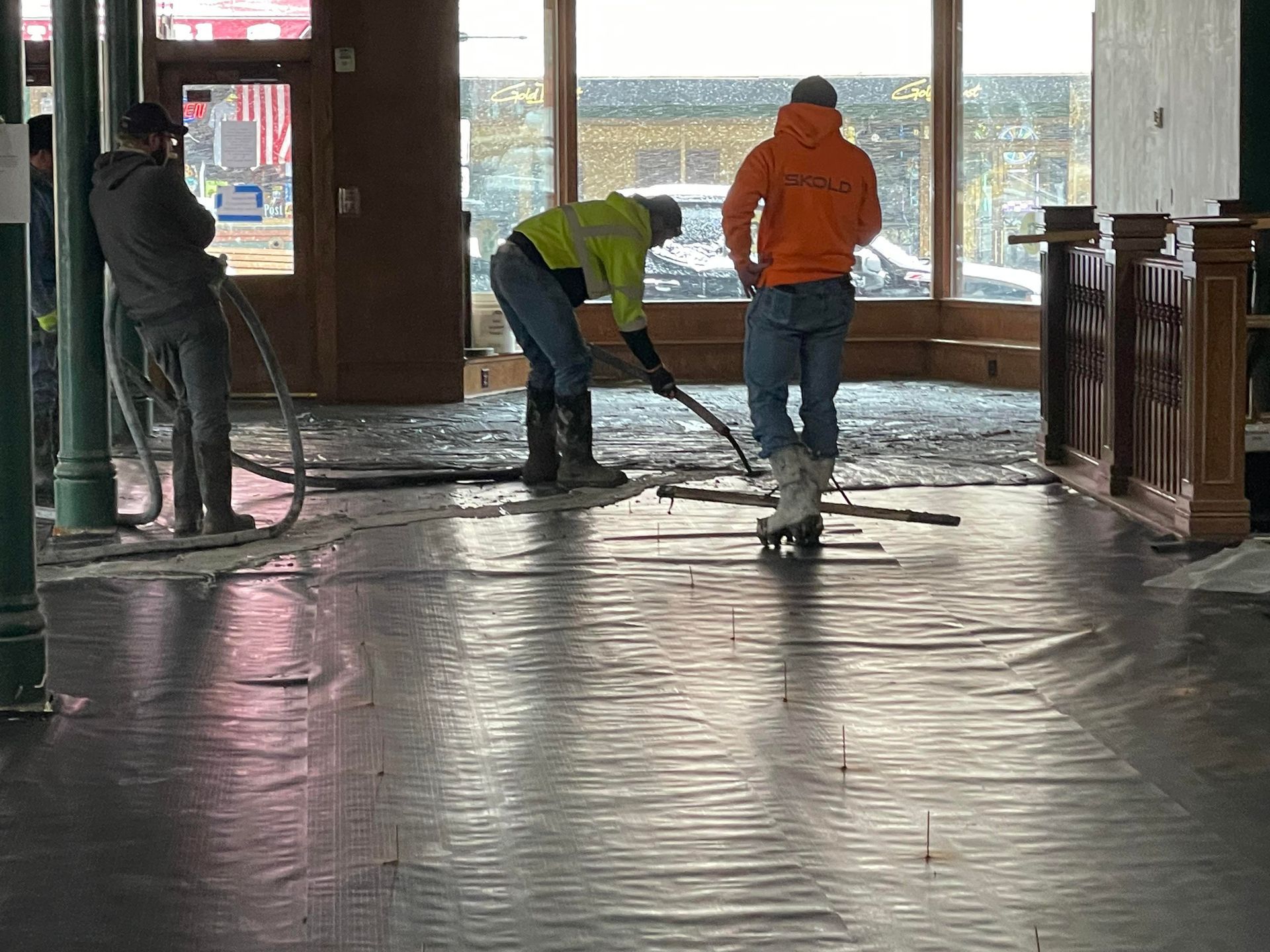 Construction workers inside a building. One uses a tool on flooring, others watch.