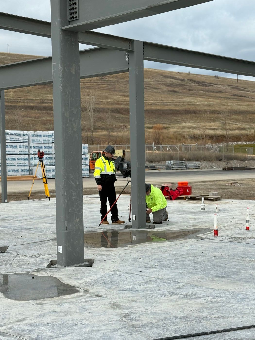 Construction workers inspect steel beams at a building site. One kneels, the other stands with a level.
