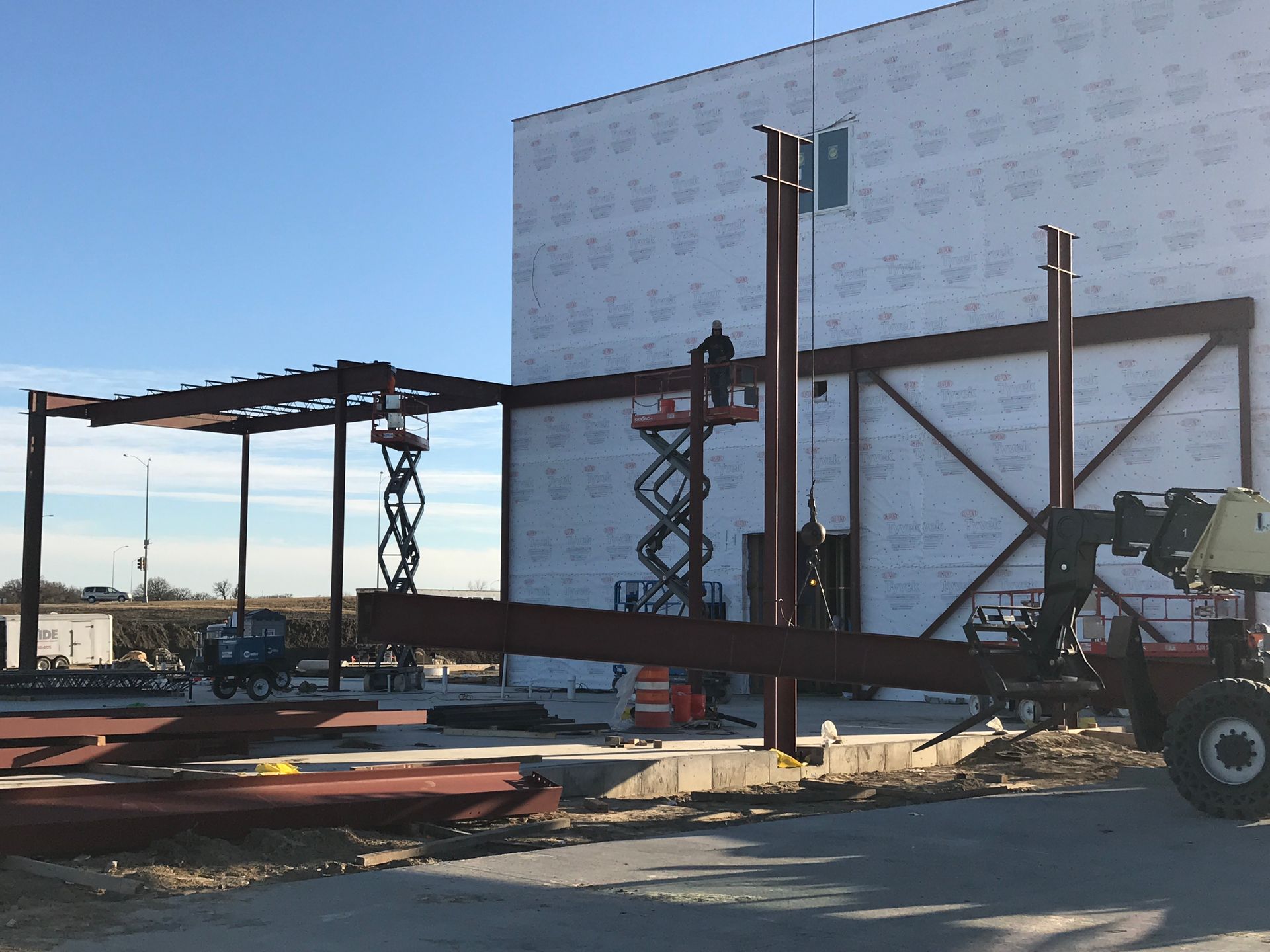 Construction site with workers, crane and beams near a building under a blue sky.