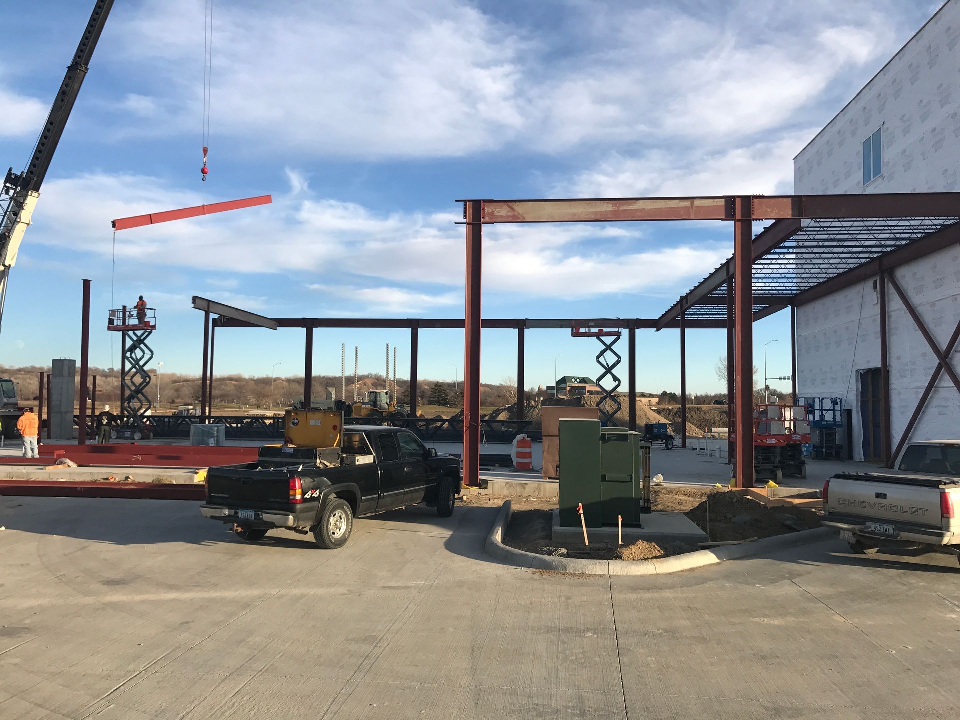 Construction site with crane lifting steel beam, workers, and trucks under a blue sky.