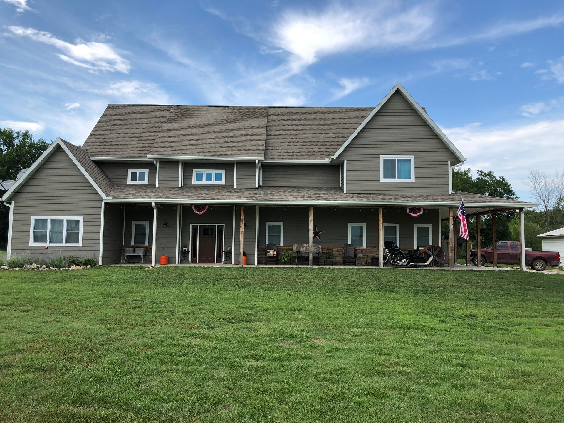 Two-story house with tan siding and a long porch, set on a green lawn under a blue sky.