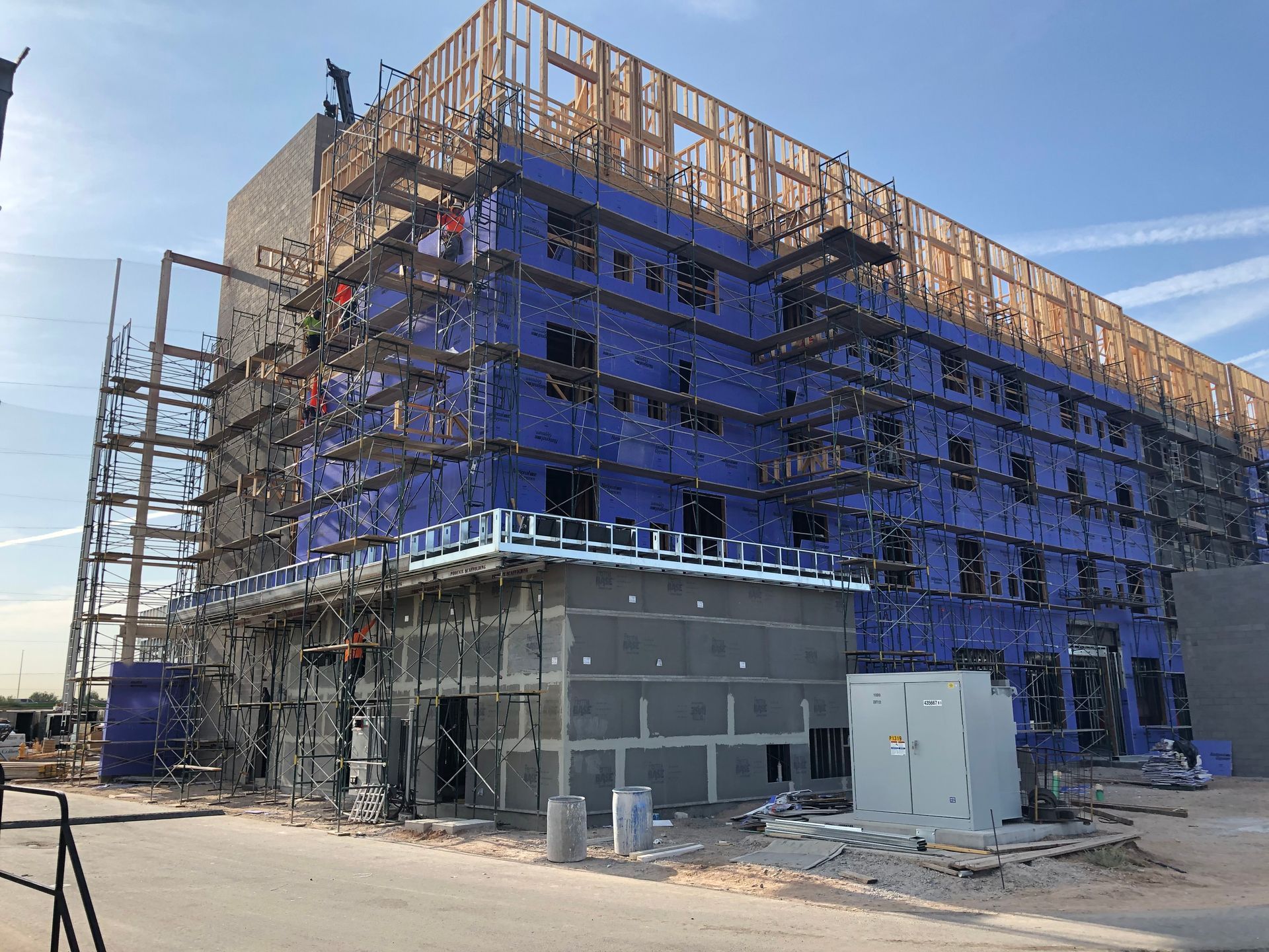 Building under construction, scaffolding and blue protective wrap visible. Wooden frame on top, daylight.