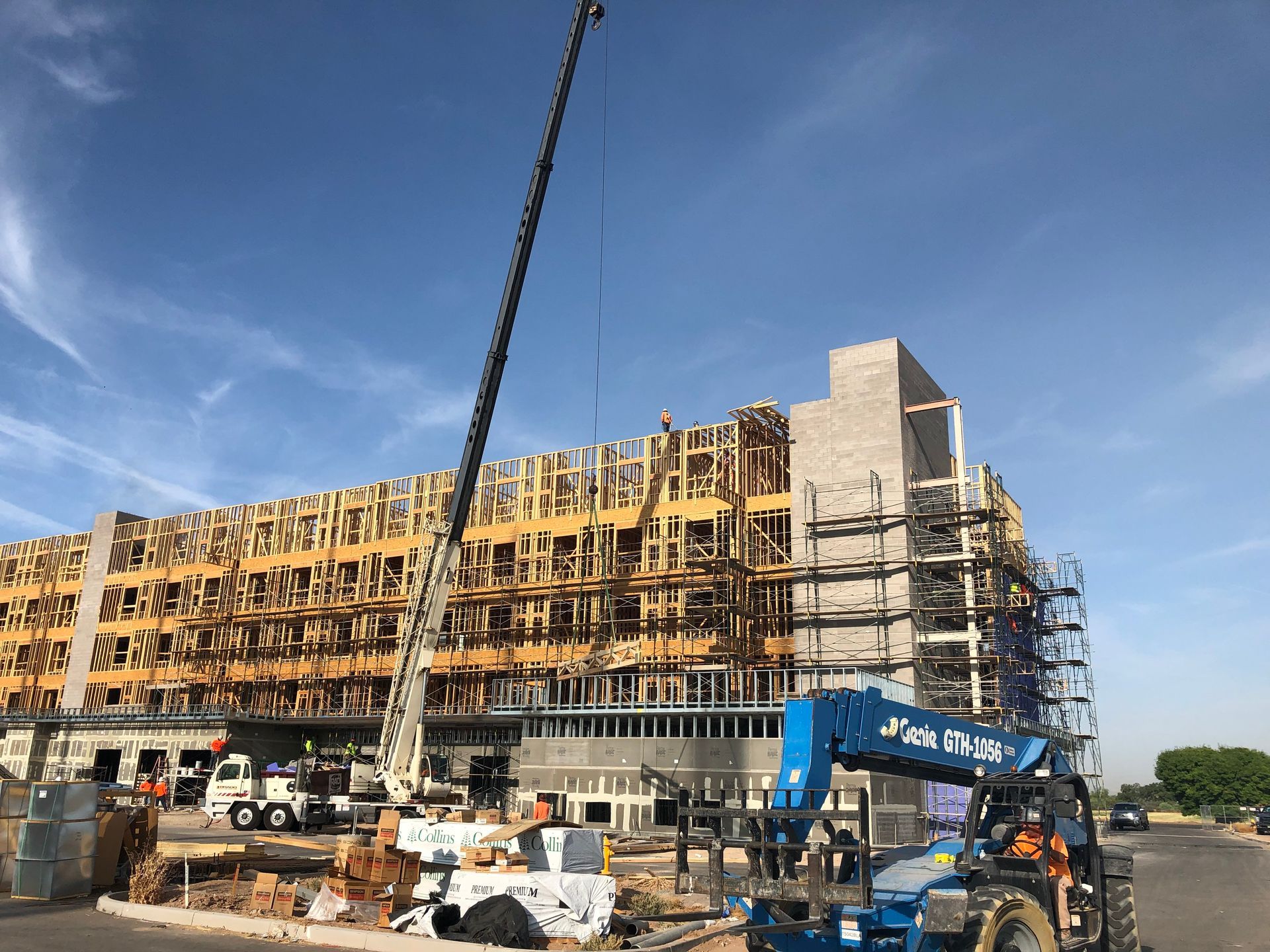 Construction site with crane lifting materials to a multi-story wooden building frame against a blue sky.