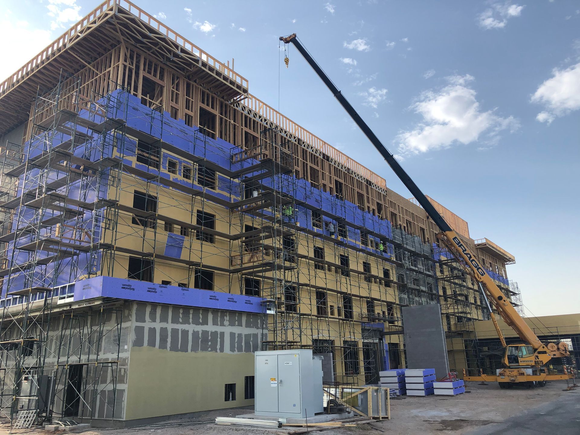 Construction site: multi-story building with wooden framing, scaffolding, crane, and insulation in progress.