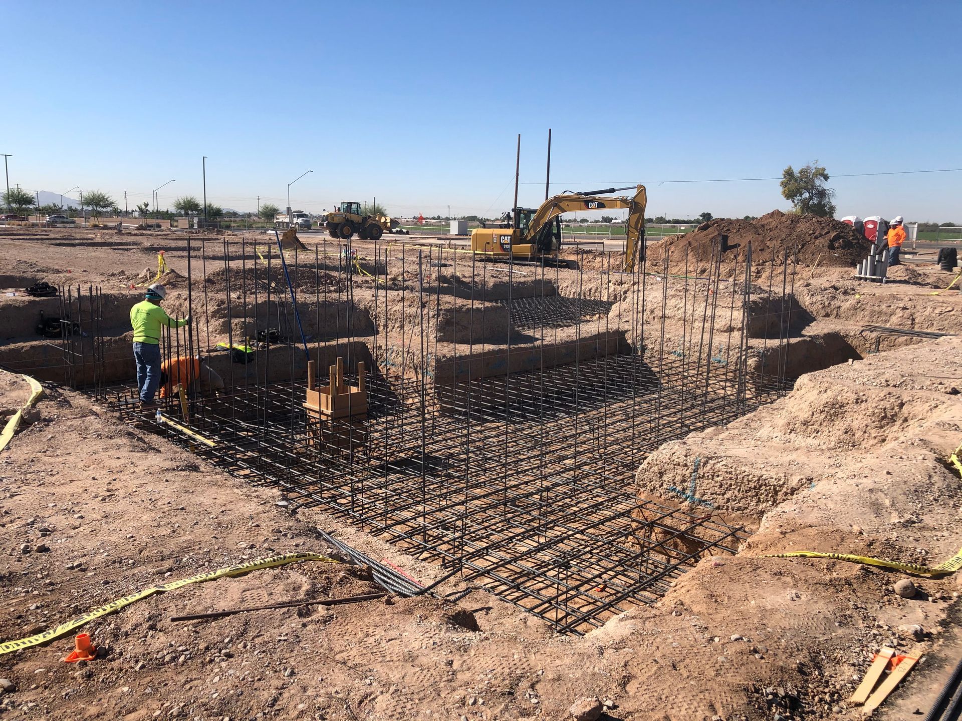 Construction site with workers, an excavator, and rebar framework in an excavated foundation.